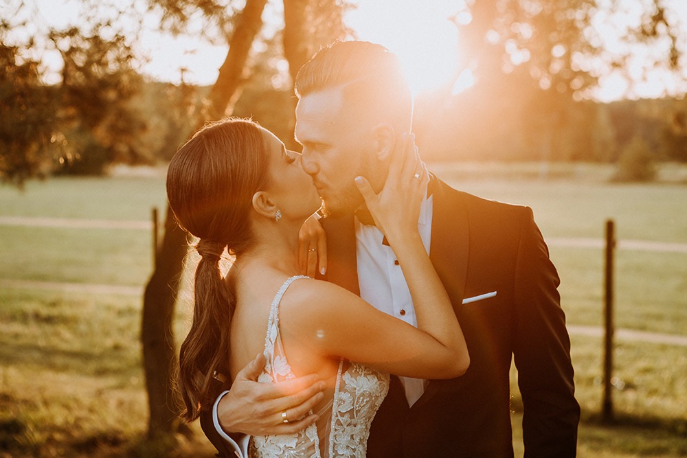 Para młoda w pocałunku, uchwycona w złotym świetle zachodzącego słońca, z widocznymi obrączkami i delikatnymi detalami sukni ślubnej.