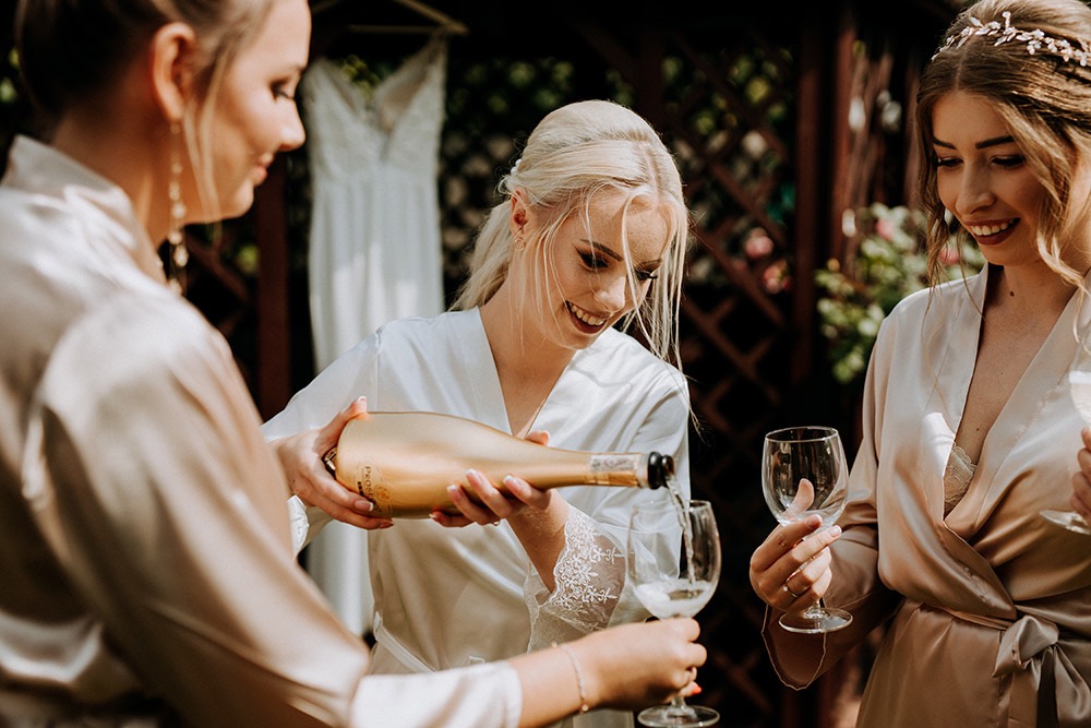 Panna młoda w białym szlafroku z koronkowymi rękawami nalewa prosecco do kieliszków dla druhen w ogrodzie, w tle suknia ślubna.