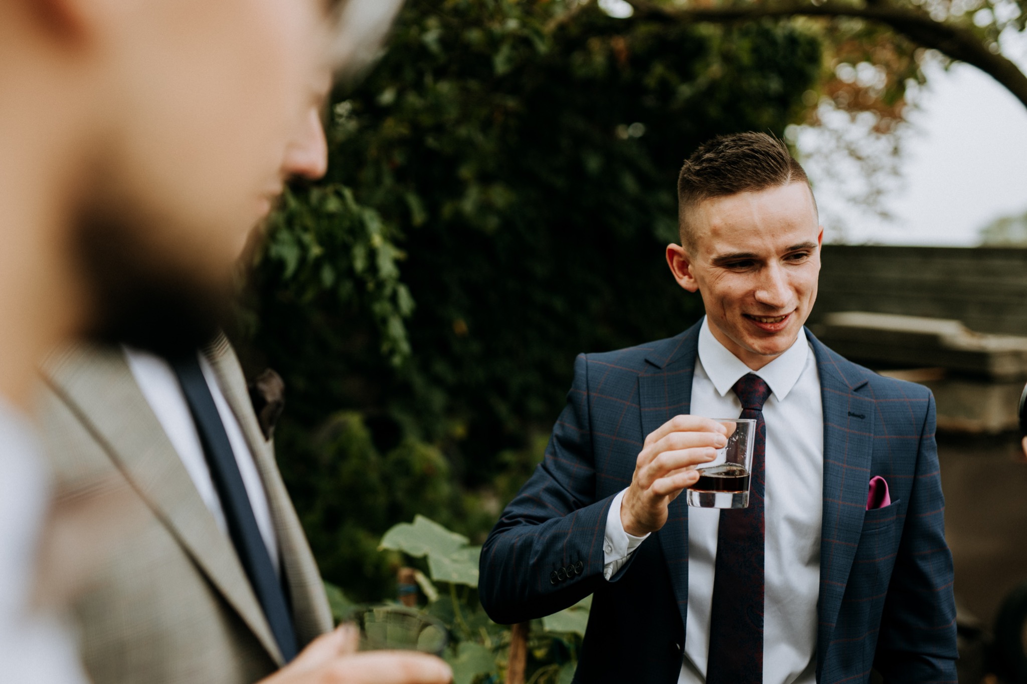 Mężczyzna w granatowym garniturze w kratę, z różową poszetką, trzyma szklankę z ciemnym napojem, uśmiechając się do osoby w beżowym garniturze po lewej stronie, na tle zieleni.