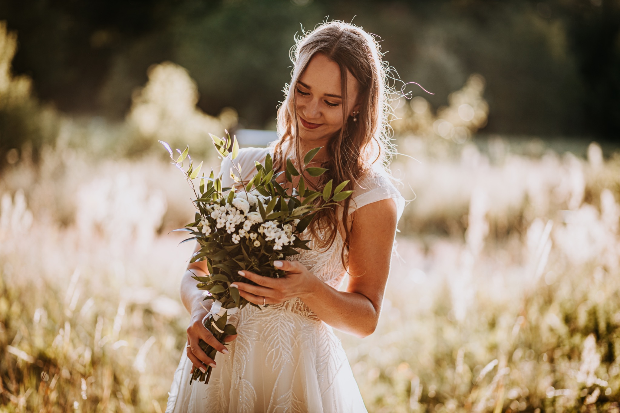 Uśmiechnięta panna młoda w białej sukni, trzymająca bukiet białych kwiatów na łące w słoneczny dzień.