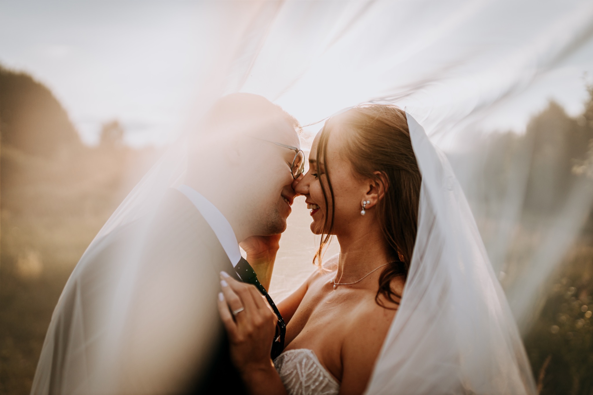 Uśmiechnięta para młoda, panna młoda z perłowymi kolczykami i delikatnym naszyjnikiem, pan młody w okularach, oboje częściowo zasłonięci welonem na tle zachodzącego słońca.