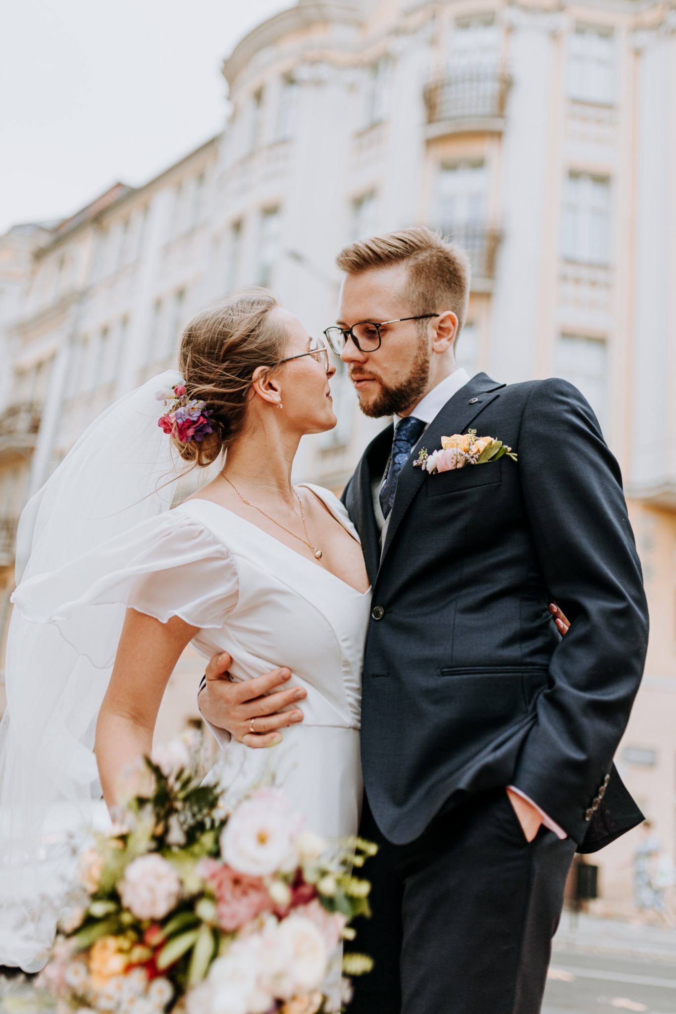 Para młoda w okularach patrzy na siebie czule na tle miejskiej architektury, panna młoda w prostej sukni z bukietem, pan młody w granatowym garniturze z butonierką.
