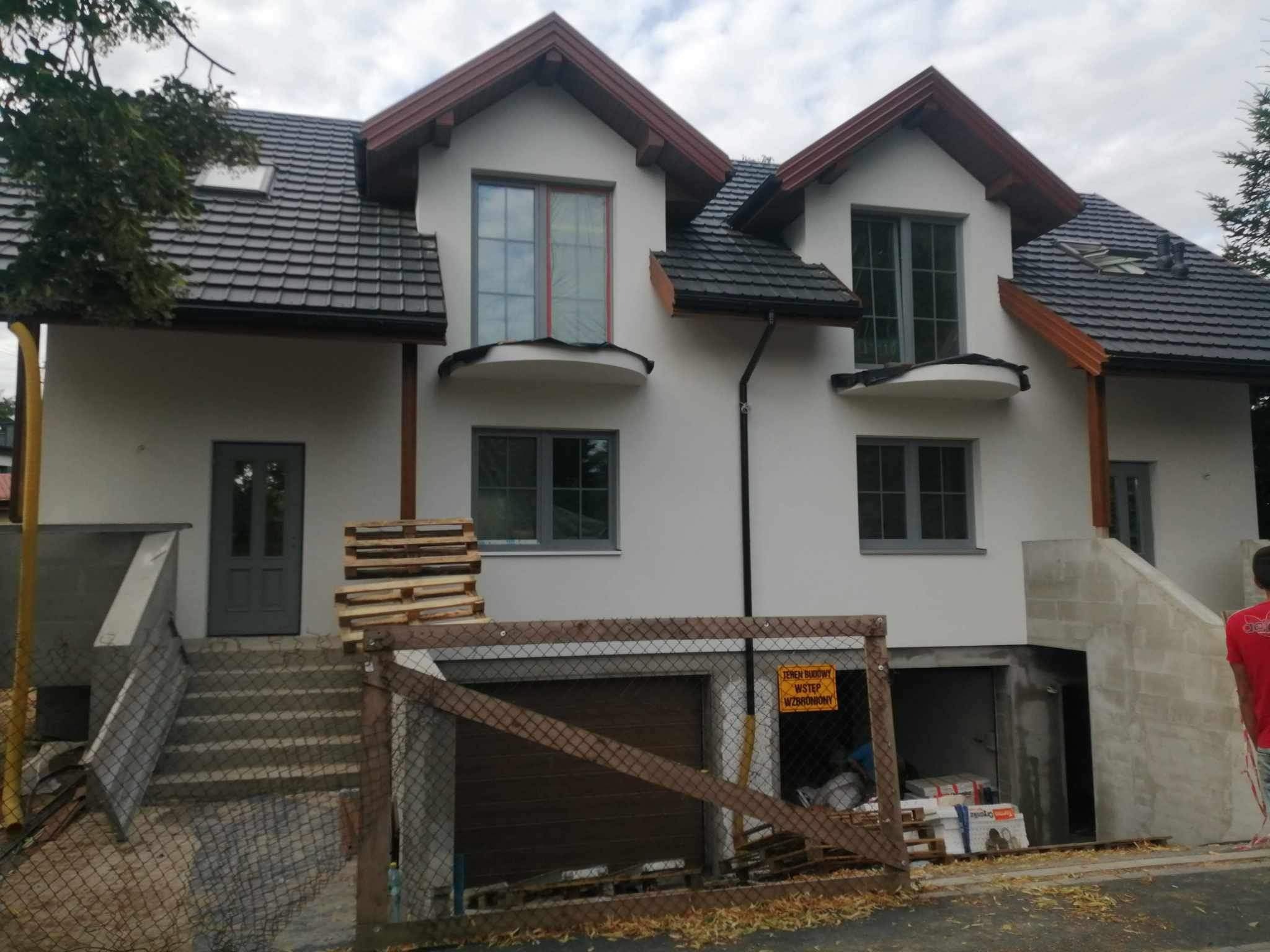 Nowo ocieplona, biała elewacja dwupiętrowego domu z szarą stolarką okienną i ciemną dachówką, widoczne drewniane palety i ogrodzenie placu budowy z żółtą tabliczką ostrzegawczą.