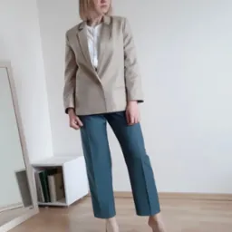 Kobieta w beżowym żakiecie, białej bluzce i turkusowych spodniach, pozująca w minimalistycznym wnętrzu z drewnianą podłogą i białymi ścianami, obok lustra i niskiej szafki z książkami.