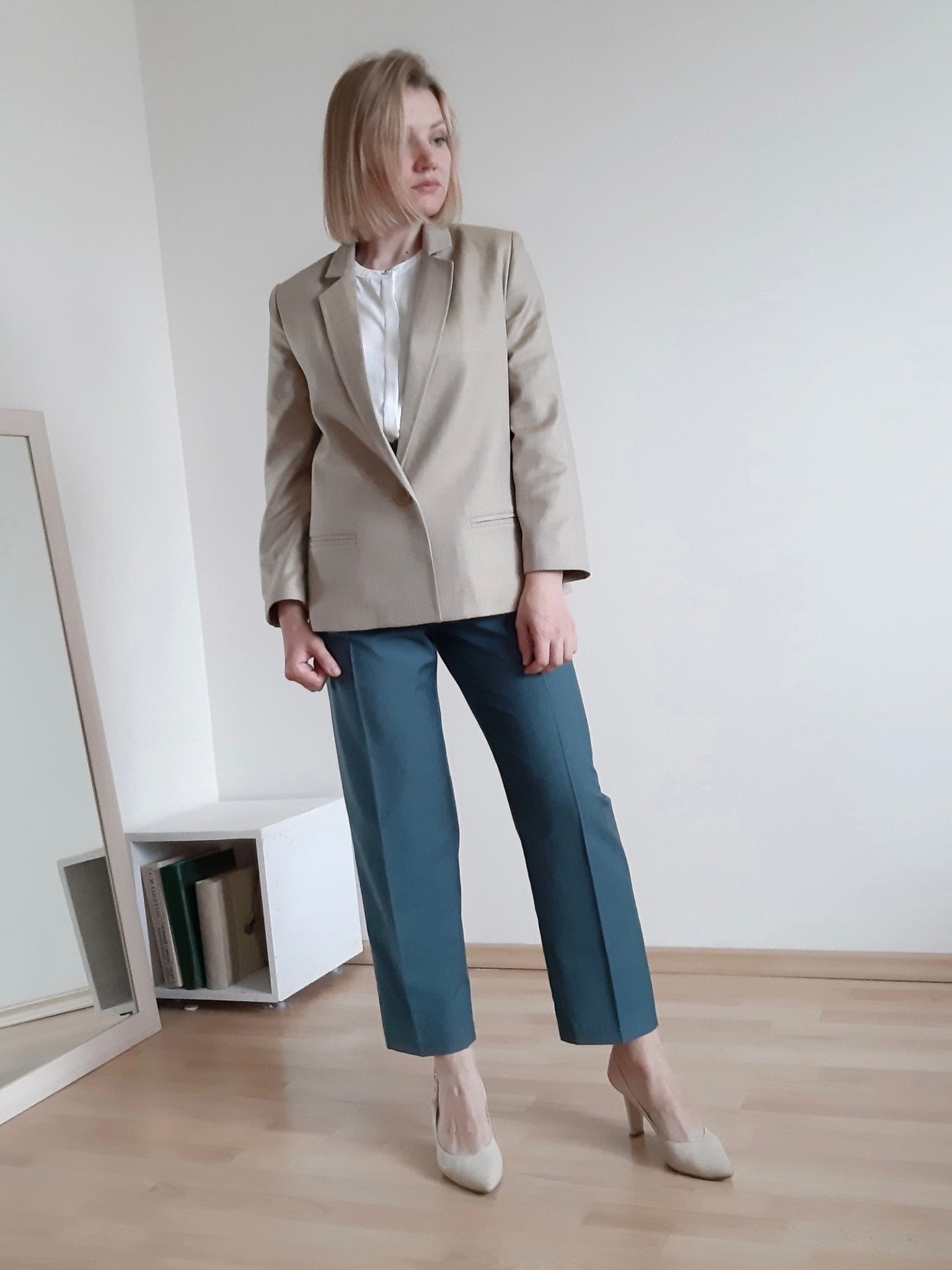 Kobieta w beżowym żakiecie, białej bluzce i turkusowych spodniach, pozująca w minimalistycznym wnętrzu z drewnianą podłogą i białymi ścianami, obok lustra i niskiej szafki z książkami.