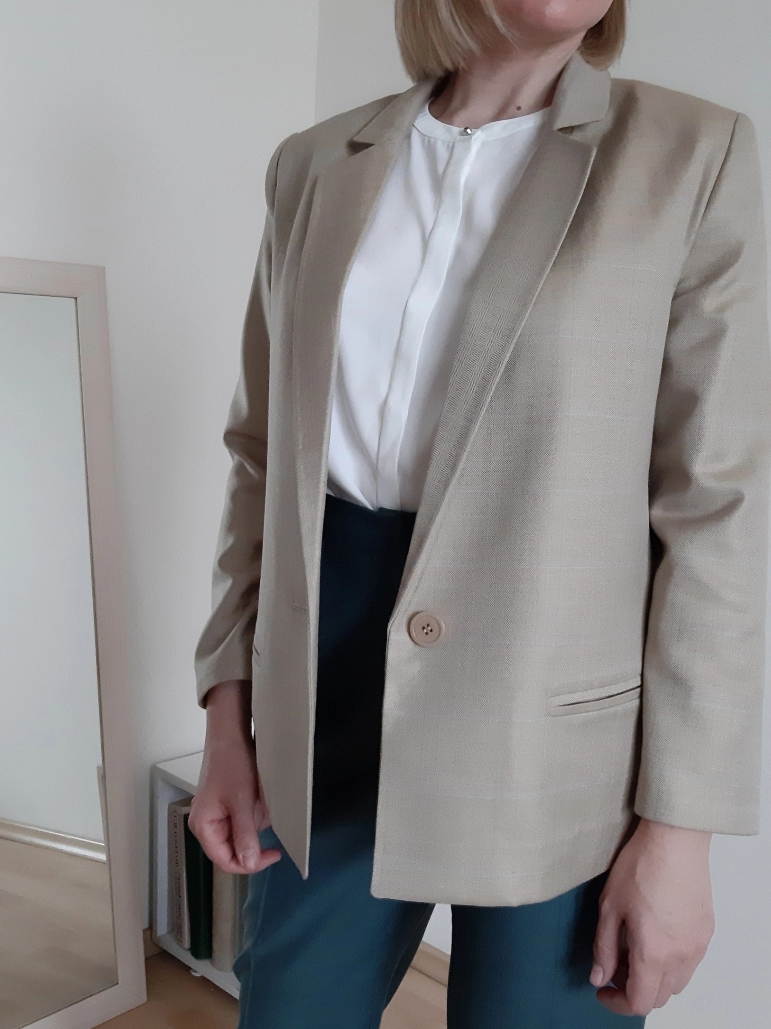 Kobieta w beżowej marynarce z pojedynczym guzikiem, białej bluzce i ciemnozielonej spódnicy, widoczna częściowo w lustrze, na tle białej ściany.
