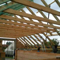 Wznoszenie drewnianej konstrukcji dachu nad budynkiem z czerwonej cegły dziurawki, widoczni pracownicy i drabina, budowa w toku.