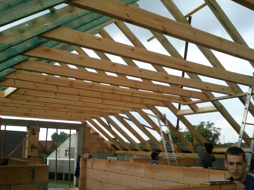 Wznoszenie drewnianej konstrukcji dachu nad budynkiem z czerwonej cegły dziurawki, widoczni pracownicy i drabina, budowa w toku.