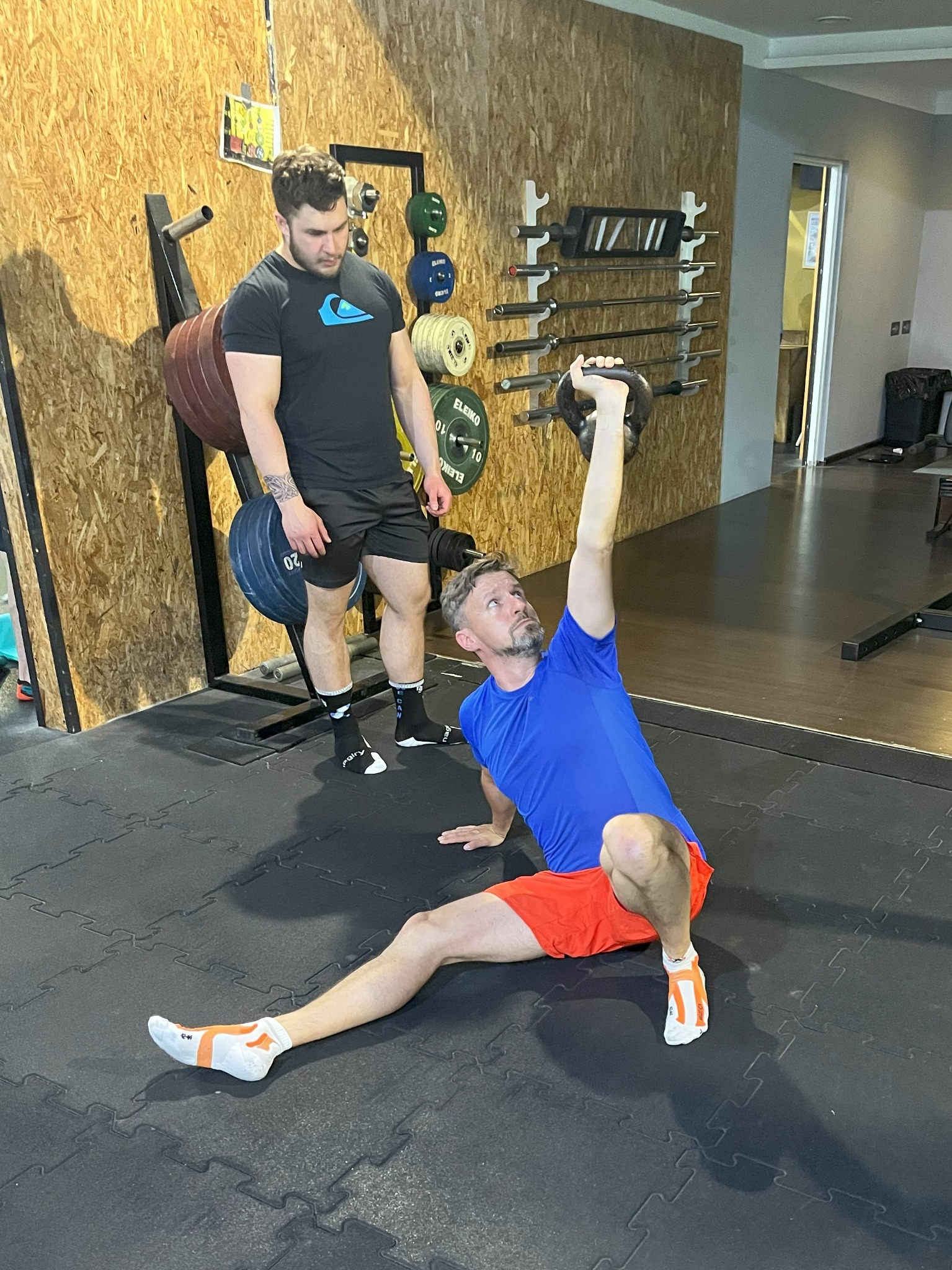 Mężczyzna w niebieskiej koszulce i pomarańczowych spodenkach wykonuje ćwiczenie z kettlebellem w siłowni, pod nadzorem trenera w czarnej koszulce, na tle drewnianej ściany z uchwytami na sztangi...