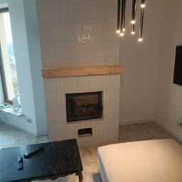 Grzegorz Ostrowski - Nowoczesny kominek obudowany białymi, wąskimi płytkami, z drewnianą półką, oświetlony wiszącymi lampami w czarnych tubach, widok z perspektywy salonu z marmurowym stolikiem i beżową sofą.