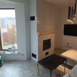 Grzegorz Ostrowski - Wykończony kominek obłożony białymi, wąskimi płytkami, z drewnianą półką, w nowoczesnym salonie z dużym oknem i czarnym stolikiem kawowym.