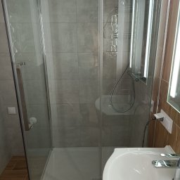 Grzegorz Ostrowski - Szklana kabina prysznicowa z brodzikiem, umywalka z baterią oraz podświetlane lustro w nowocześnie wykończonej łazience z szarymi płytkami.