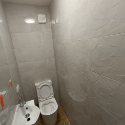 Grzegorz Ostrowski - Nowoczesna łazienka po remoncie: biała umywalka z szafką, toaleta z otwartą klapą, szare płytki z geometrycznym wzorem na ścianach, drewnopodobna podłoga, wentylator sufitowy.