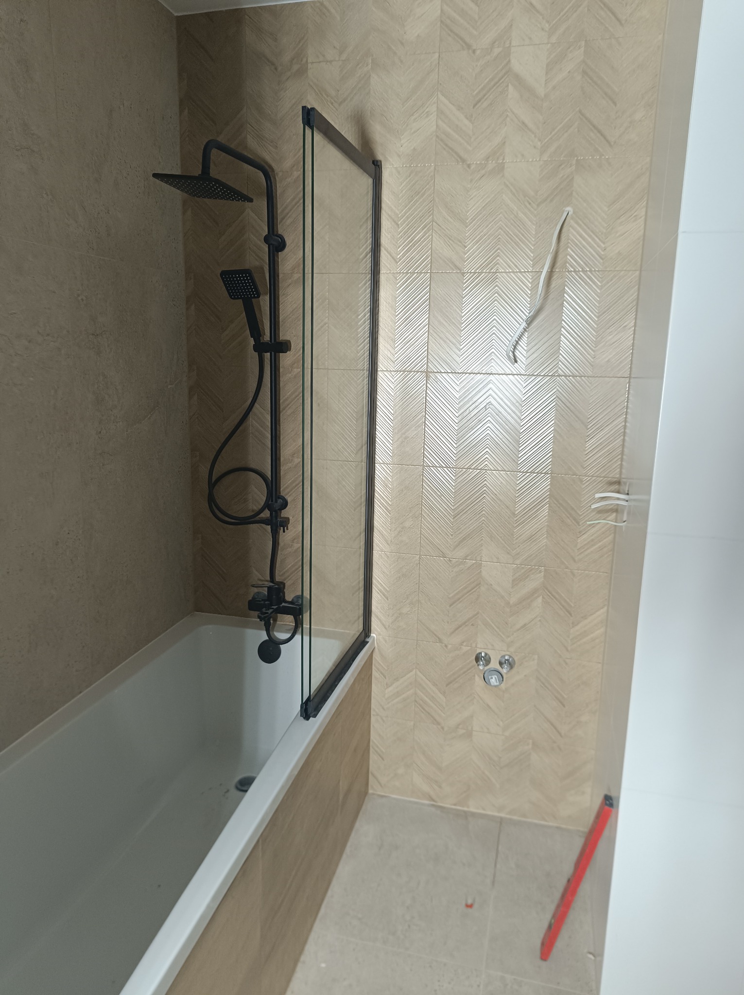 Wykończona łazienka z wanną, czarną armaturą prysznicową i szklanym parawanem nawannowym, płytki w jodełkę na ścianach, widoczny poziomica na podłodze.