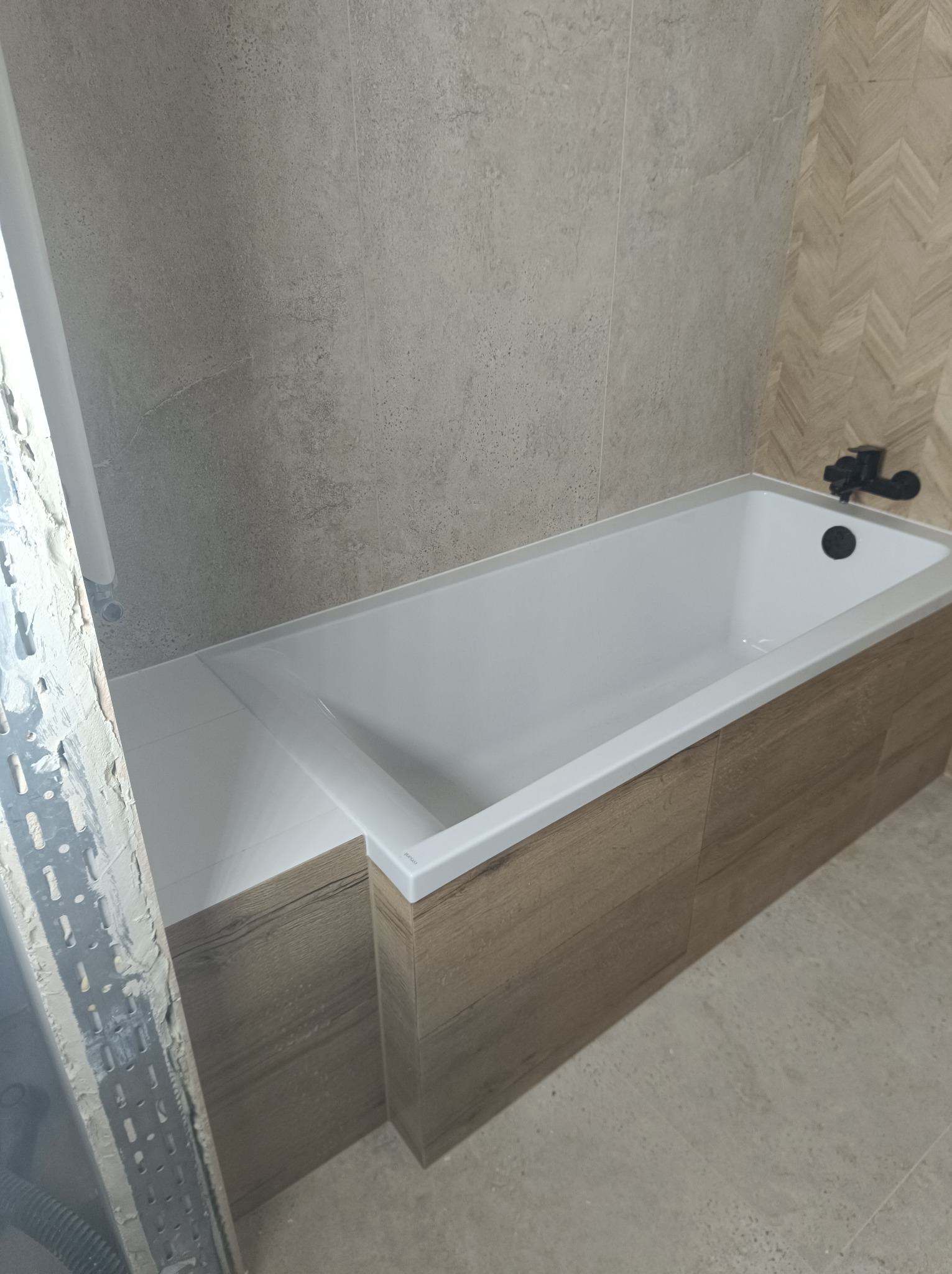 Biała wanna obudowana panelami imitującymi drewno, z czarną armaturą, na tle beżowych płytek, z widocznym fragmentem ściany w trakcie prac wykończeniowych.