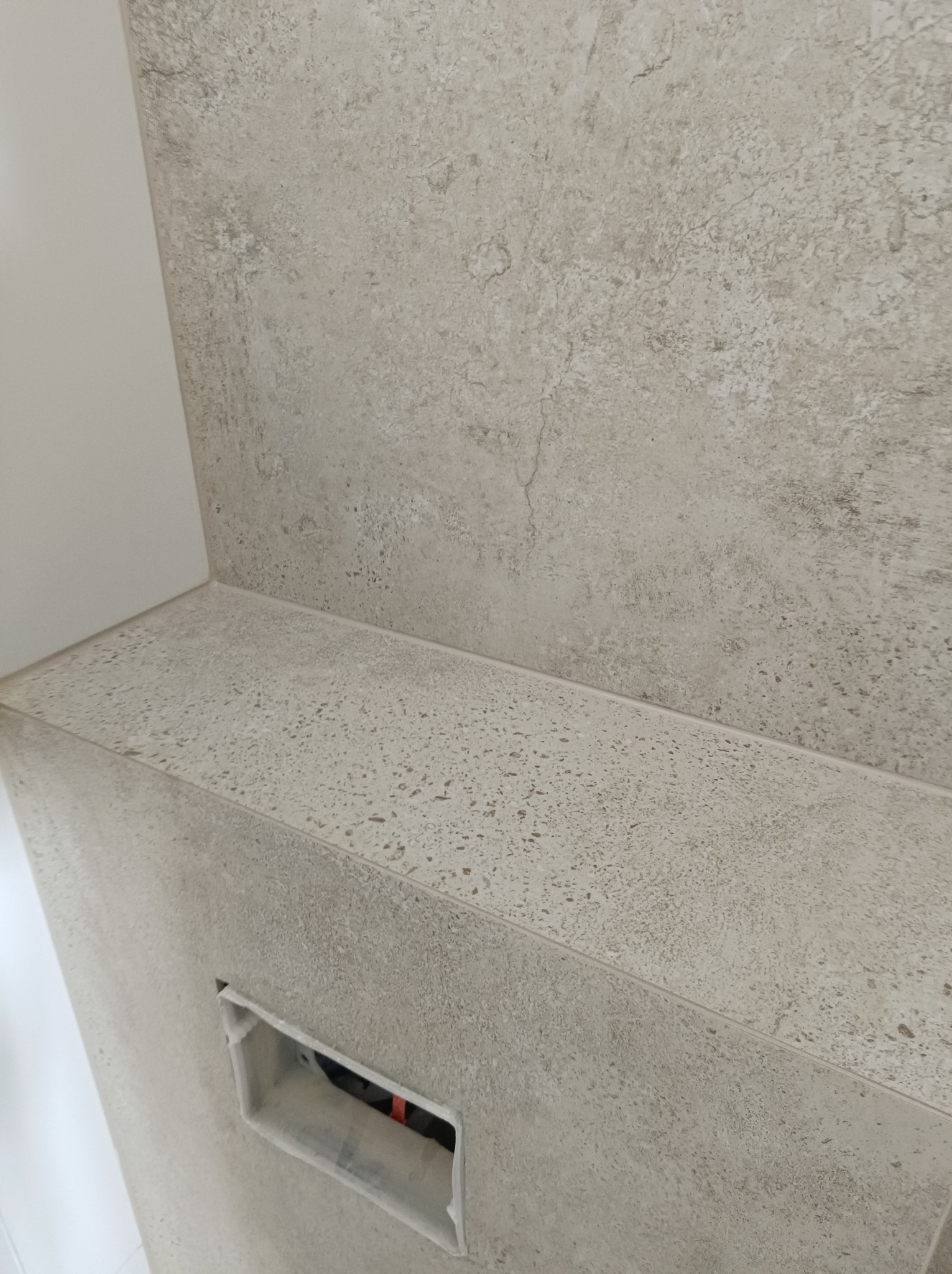 Fragment ściany i półki wykończonej płytkami imitującymi kamień w łazience, widoczny otwór na instalację.