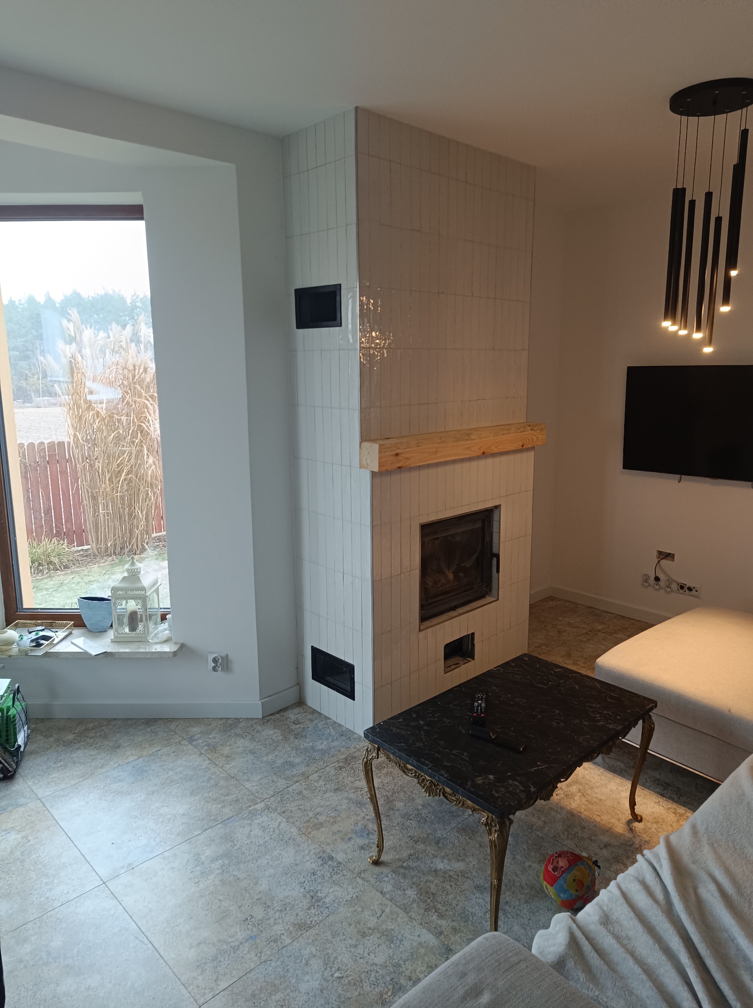 Wykończony kominek obłożony białymi, wąskimi płytkami, z drewnianą półką, w nowoczesnym salonie z dużym oknem i czarnym stolikiem kawowym.