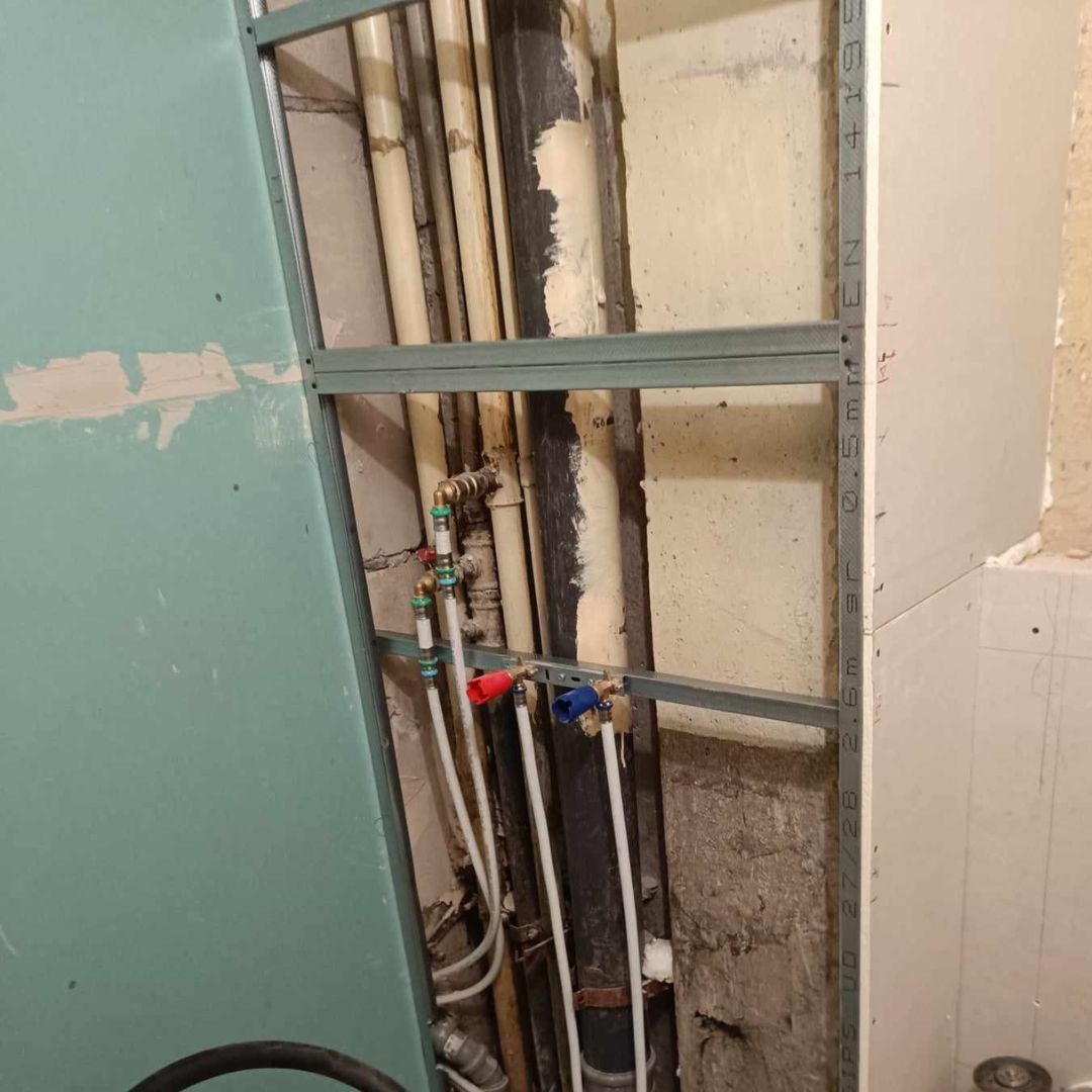 Fragment ściany podczas prac instalacyjnych: widoczne rury wodne z zaworami oznaczonymi kolorami, ramy pod płyty gipsowo-kartonowe i fragment ściany z widocznymi warstwami wykończeniowymi.