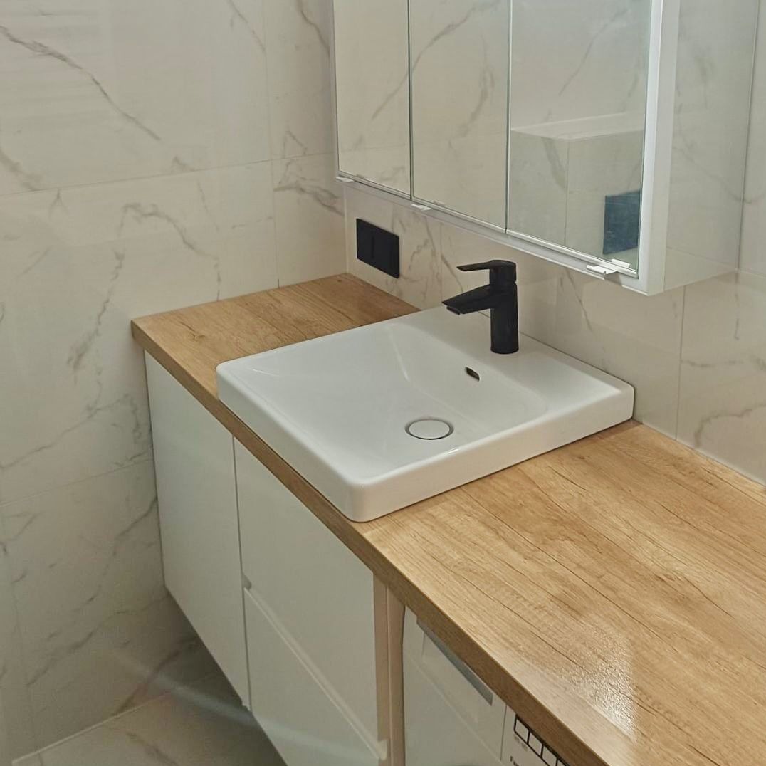 Biała, prostokątna umywalka nablatowa z czarną, minimalistyczną baterią na blacie imitującym drewno, z szafką pod umywalką i lustrem w szafce wiszącej, białe płytki marmurowe w tle.