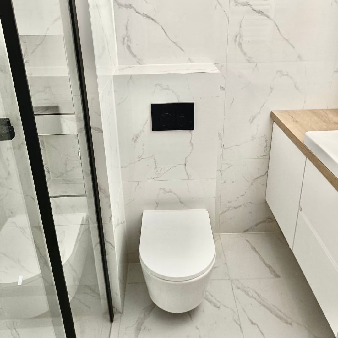 Nowoczesna łazienka z białymi marmurkowymi płytkami, wiszącą toaletą z czarnym przyciskiem spłukującym, kabiną prysznicową z czarnymi ramami i białą szafką umywalkową z drewnianym blatem.