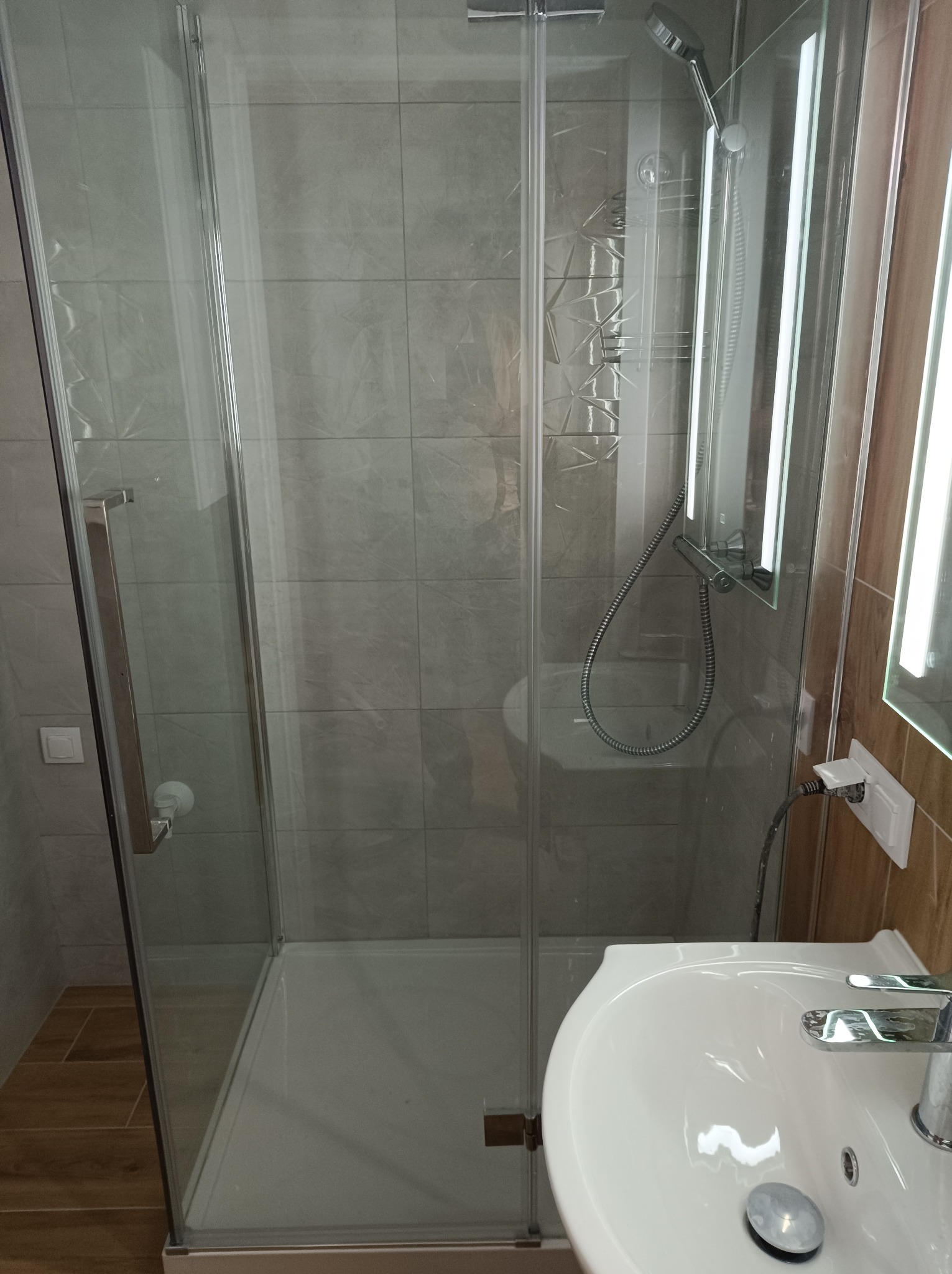 Szklana kabina prysznicowa z brodzikiem, umywalka z baterią oraz podświetlane lustro w nowocześnie wykończonej łazience z szarymi płytkami.