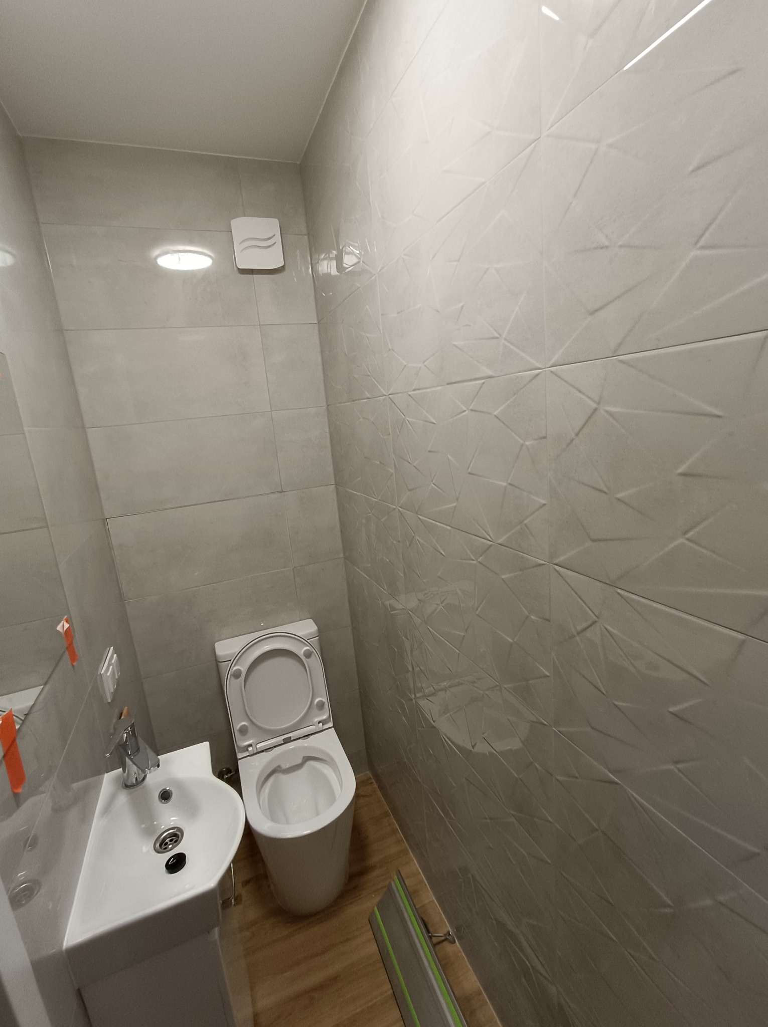 Nowoczesna łazienka po remoncie: biała umywalka z szafką, toaleta z otwartą klapą, szare płytki z geometrycznym wzorem na ścianach, drewnopodobna podłoga, wentylator sufitowy.