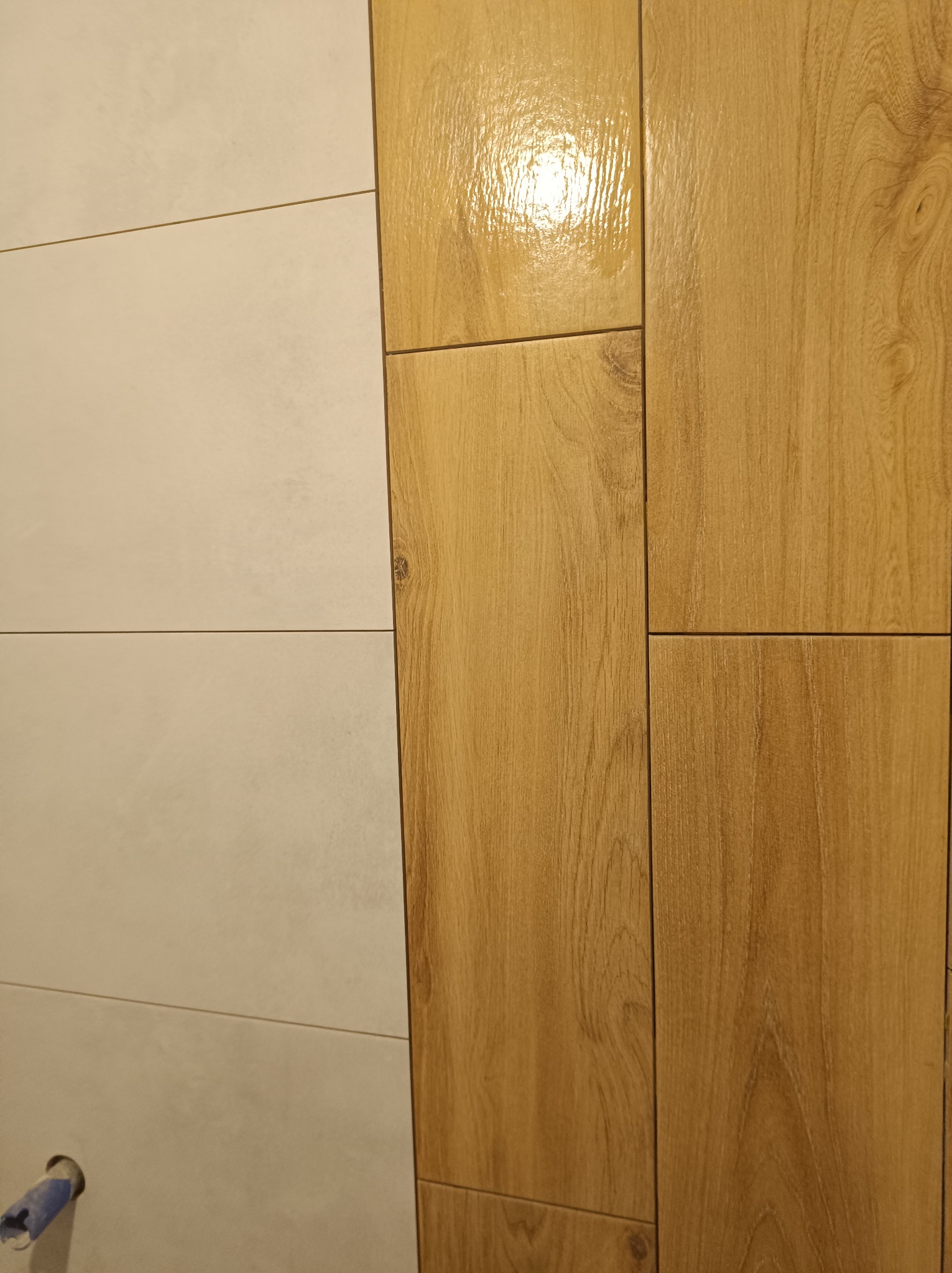 Fragment ściany wyłożonej jasnymi, prostokątnymi płytkami ceramicznymi sąsiadującymi z pionowym pasem płytek imitujących drewno o ciepłym odcieniu, widoczny otwór na rurę.