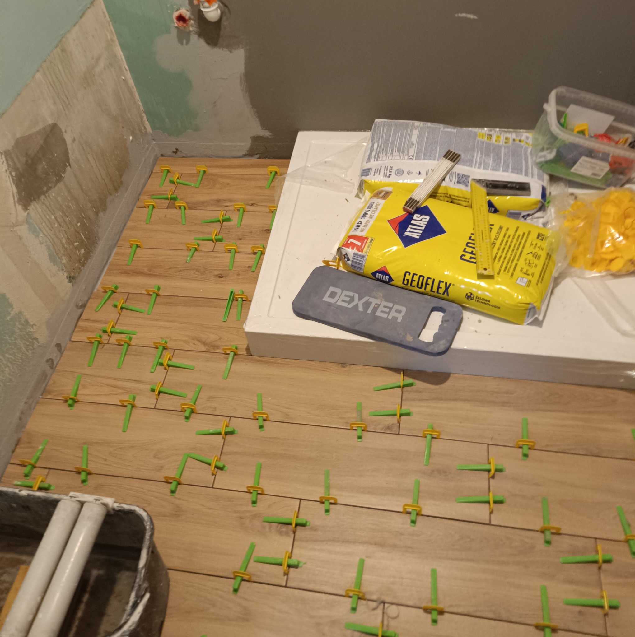 Ułożone płytki podłogowe imitujące drewno, z zielonymi krzyżykami dystansowymi, obok worek z klejem Atlas Geoflex, paczka płytek i wiadro z narzędziami w trakcie prac wykończeniowych.