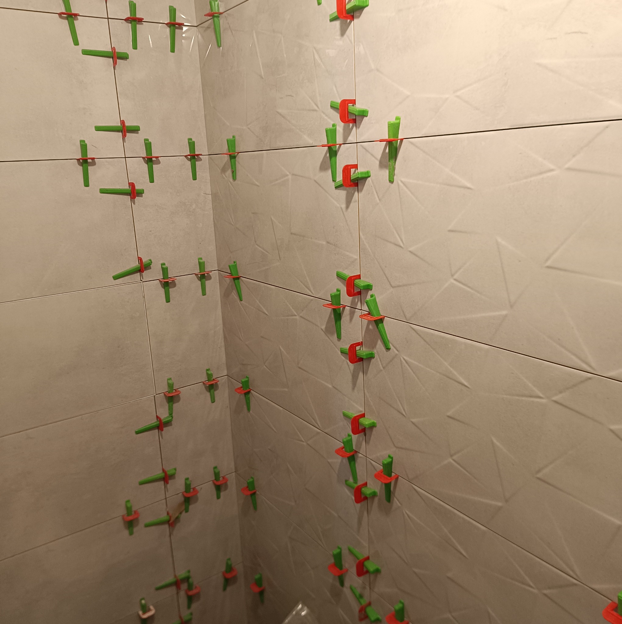 Świeżo ułożone, beżowe płytki ścienne z geometrycznym wzorem, zabezpieczone systemem poziomowania z zielonymi klinami i czerwonymi klipsami dystansowymi na czarnej lince poziomującej.