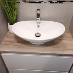 Biała, owalna umywalka nablatowa z chromowaną baterią i korkiem automatycznym, ustawiona na szafce z drewnopodobnym blatem. Obok, doniczka z zieloną trawą ozdobną. W tle, szara ściana...