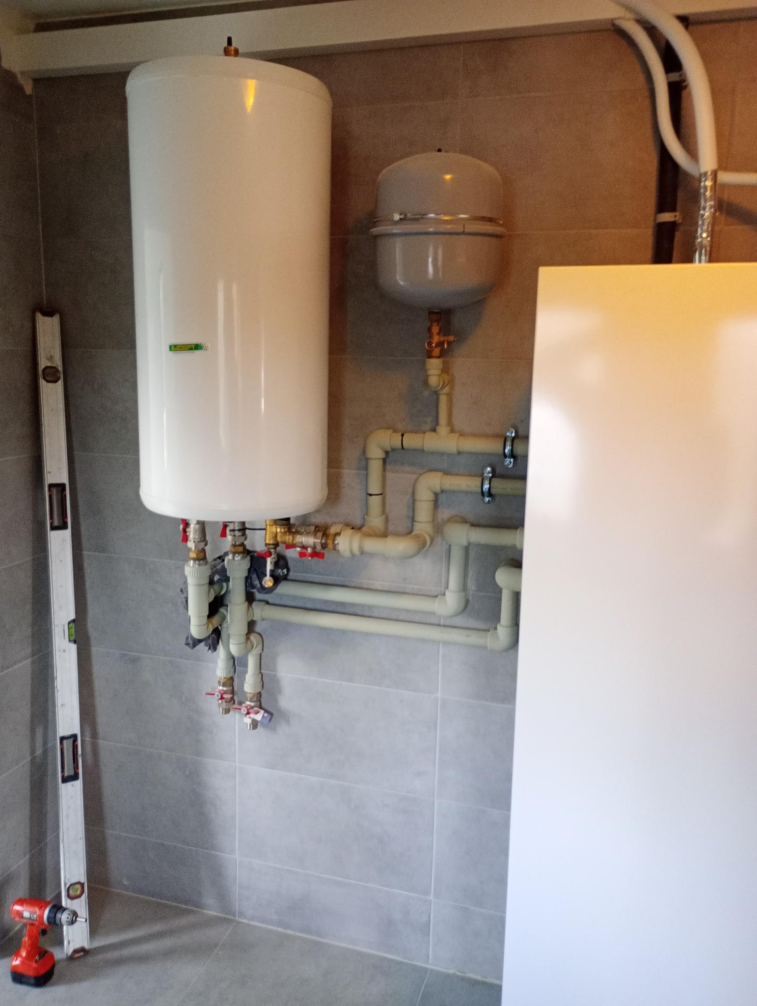 Bojler z podłączonymi rurami i osprzętem, poziomica oparta o ścianę, wiertarka leżąca na podłodze, szare płytki na ścianie i podłodze.