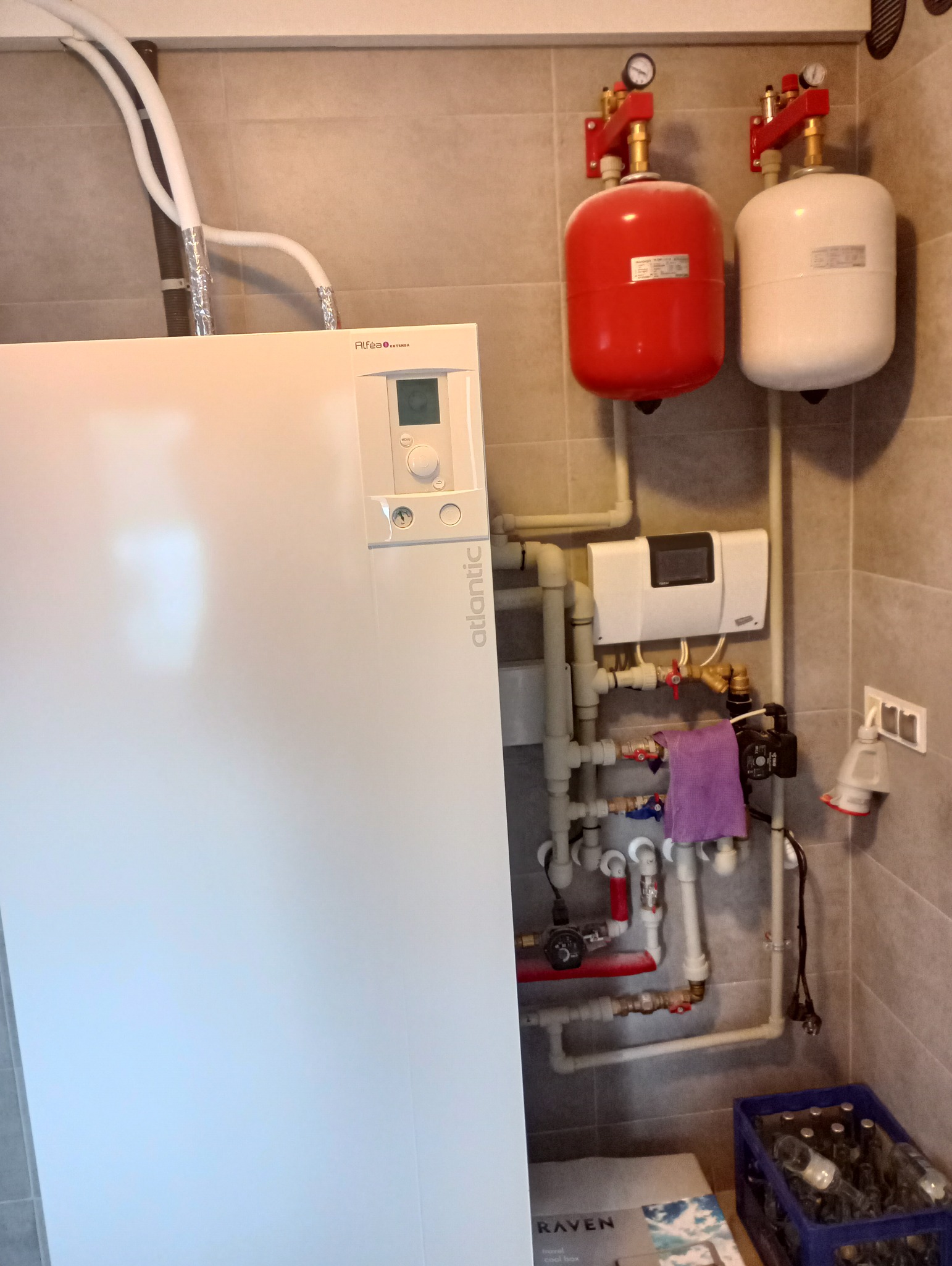 Biały piec Atlantic z podłączonymi rurami, czerwony i biały zbiornik wyrównawczy z manometrami, instalacja hydrauliczna z pompą obiegową i fioletową ścierką, niebieski koszyk z butelkami na podłodze.