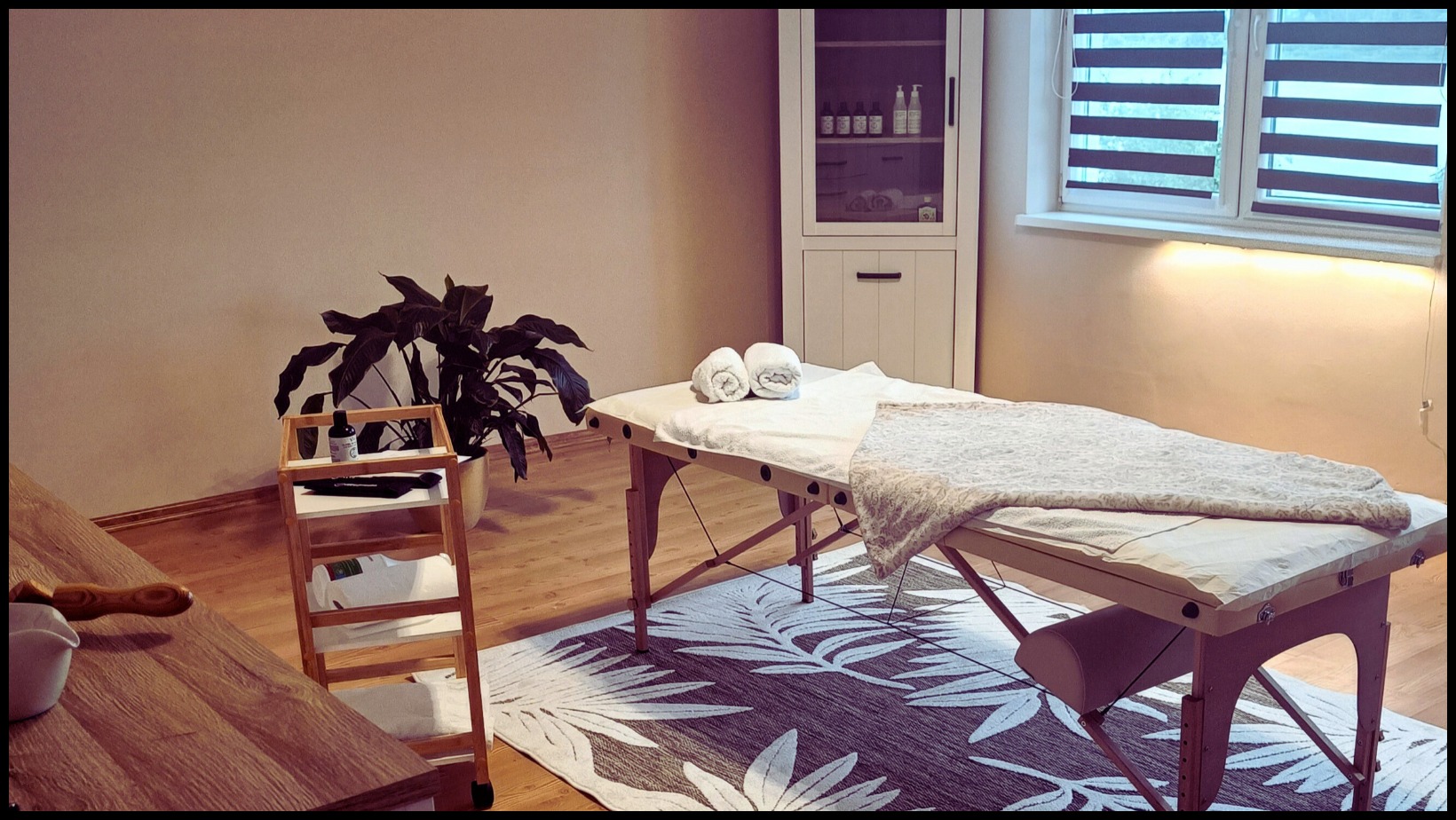 Przytulne wnętrze gabinetu z drewnianym łóżkiem do zabiegów, zwinięte ręczniki, dekoracyjny pled, roślina w doniczce na wózku i szafka z kosmetykami w tle. Na podłodze dywan z motywem liści.