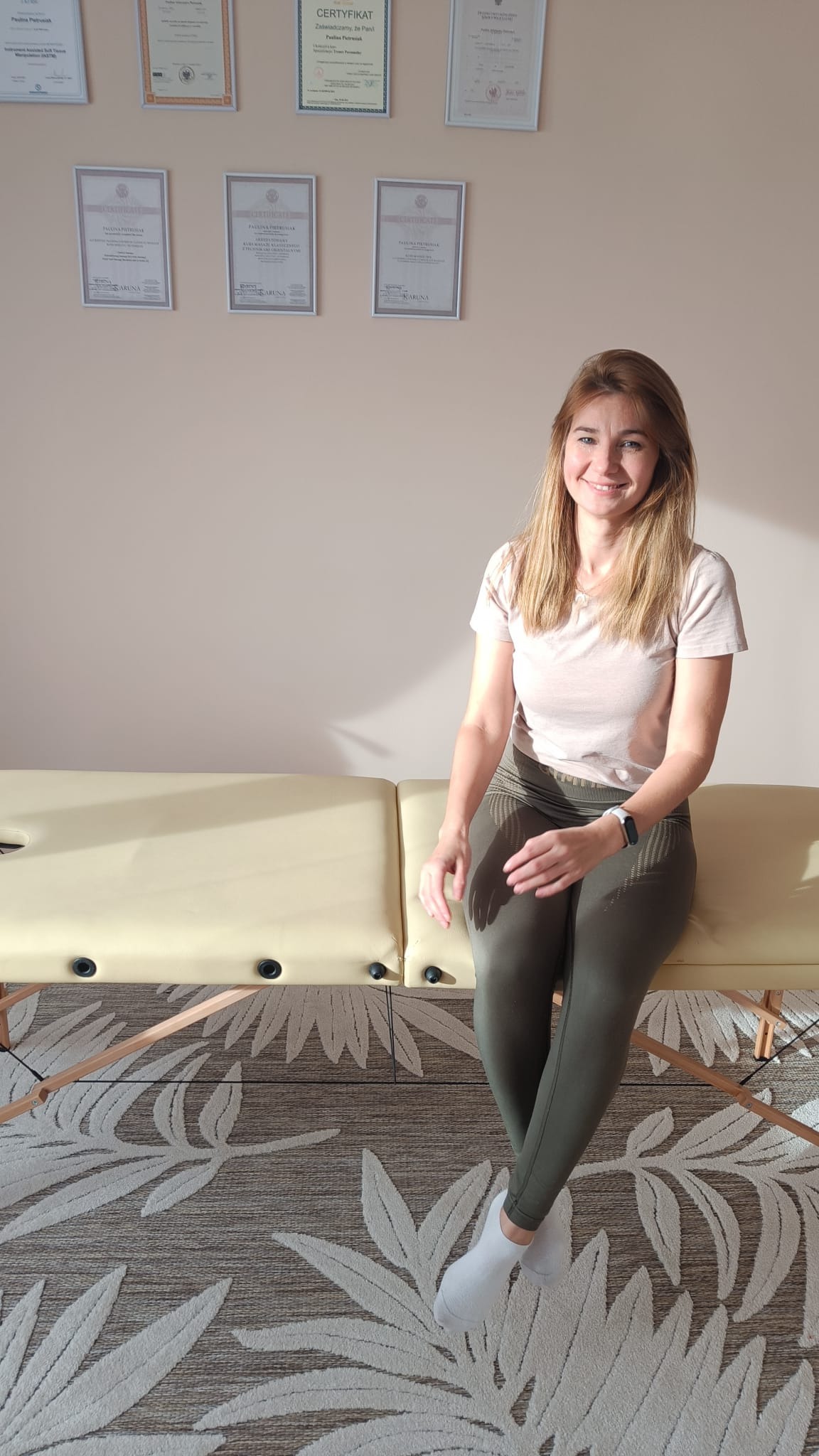 Uśmiechnięta kobieta siedzi na beżowym stole do zabiegów, na ścianie wiszą certyfikaty w ramkach, a na podłodze leży dywan w liście.