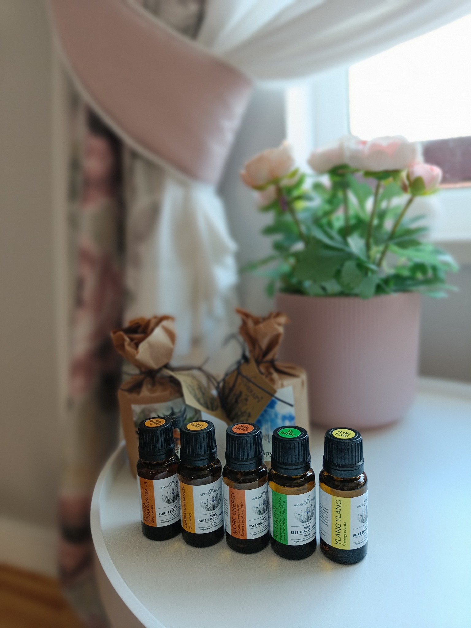 Pięć buteleczek olejków eterycznych AromatherapyOils z etykietami 'Słodka Pomarańcza', 'Gorzka Pomarańcza', 'More Energy', 'Be Happy' i 'Ylang Ylang', ustawionych na białym stoliku obok okna...