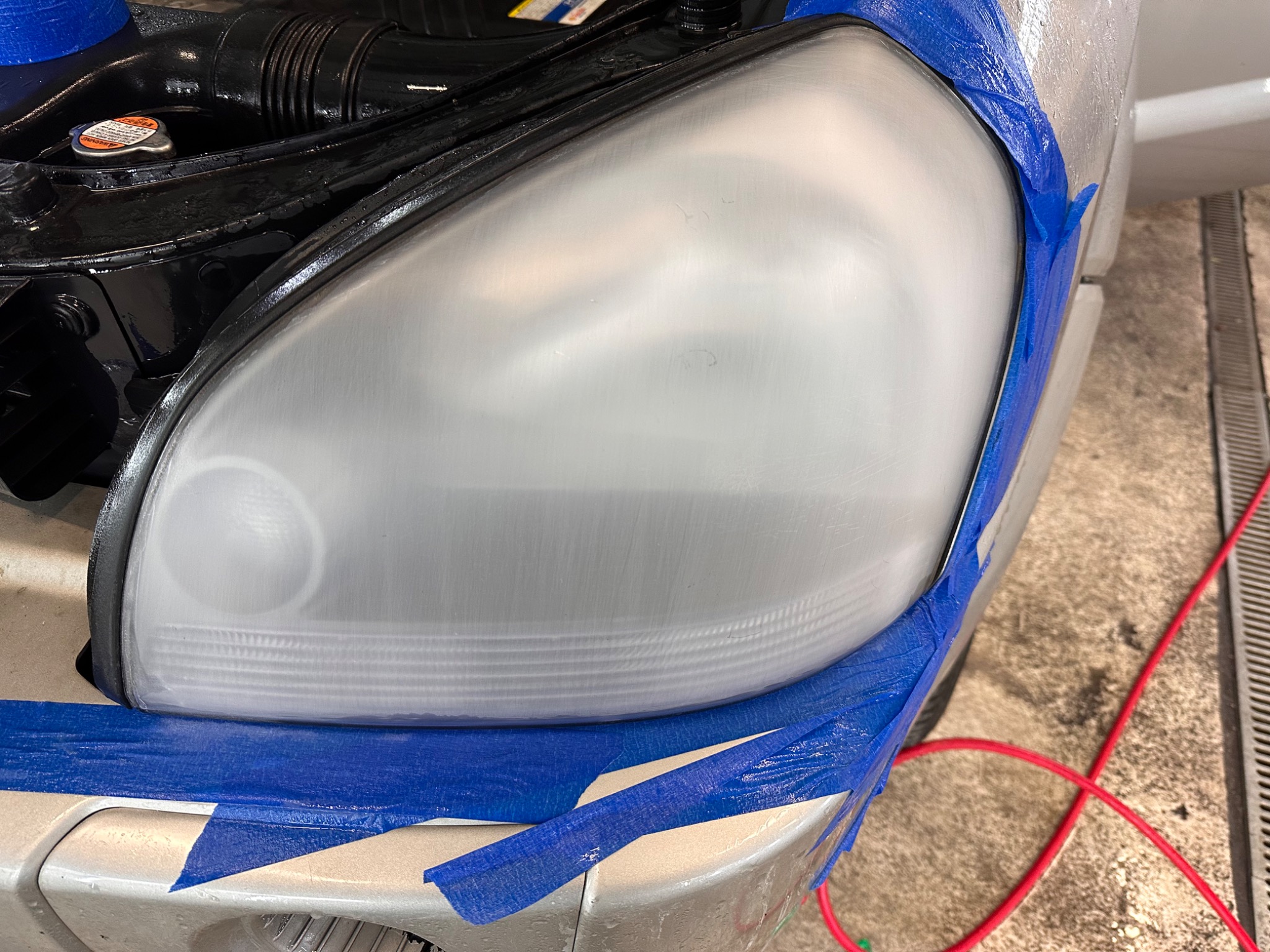 Matowa, zarysowana lampa samochodowa zabezpieczona niebieską taśmą malarską przed renowacją, widoczny fragment silnika i czerwony przewód.