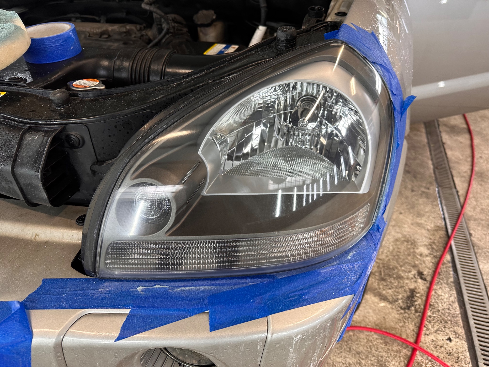 Reflektor samochodu oklejony niebieską taśmą malarską podczas renowacji, widoczne ślady polerowania.