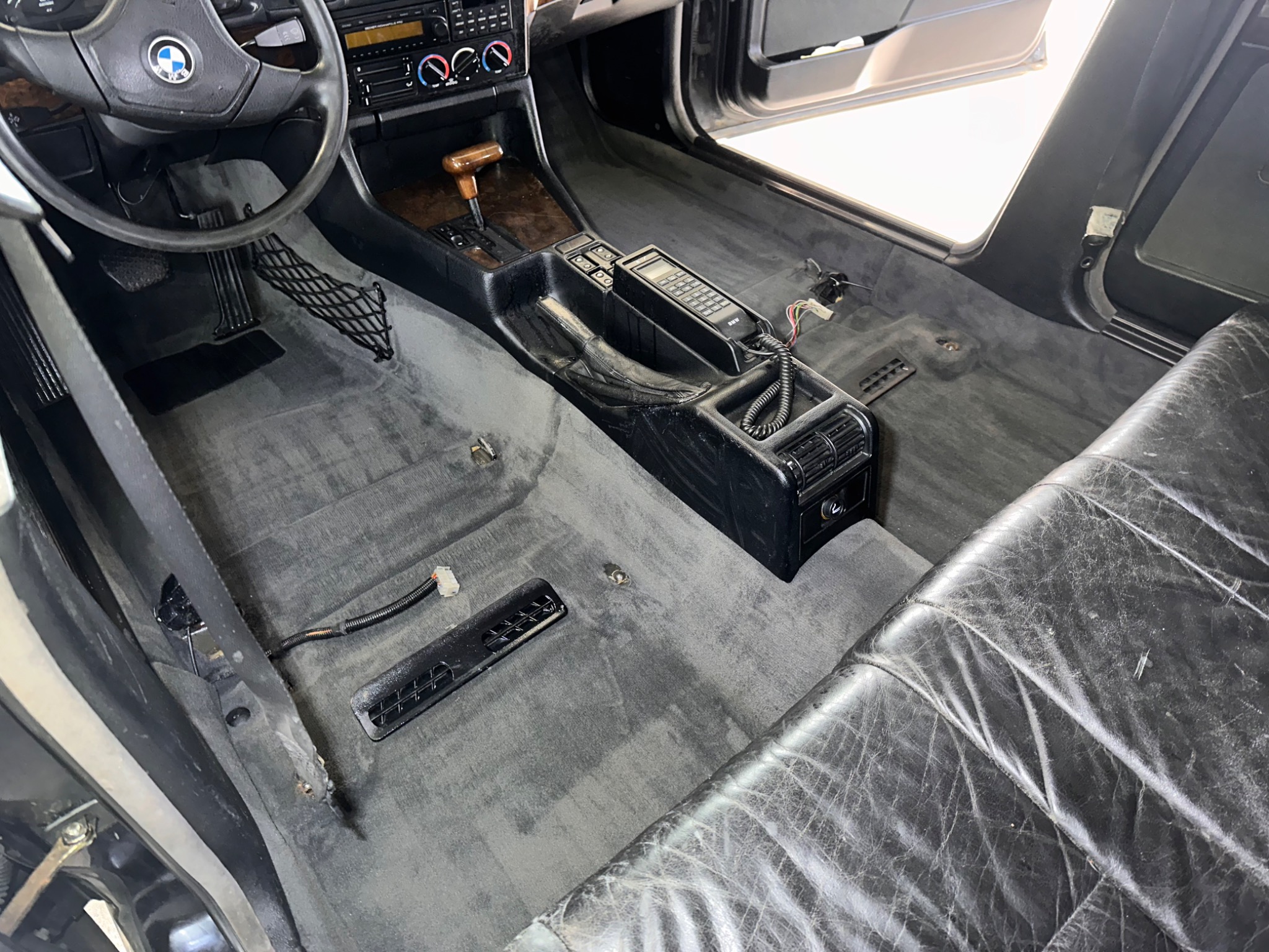 Wnętrze samochodu BMW E34 przed praniem: widoczny brudny dywanik podłogowy, skórzane fotele z widocznymi śladami użytkowania oraz oryginalny telefon samochodowy w konsoli środkowej.