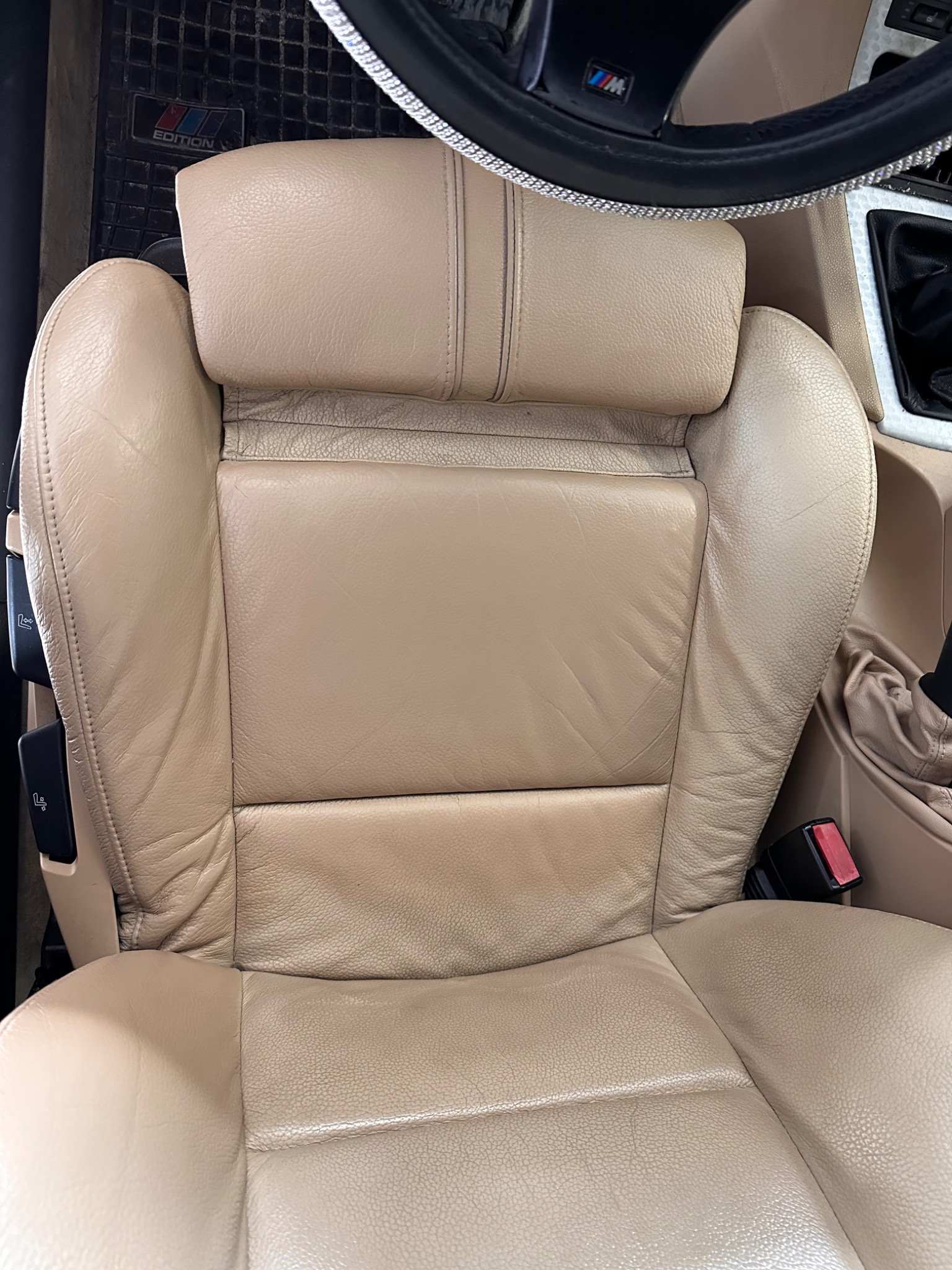 Beżowe skórzane siedzenie samochodowe z widocznymi elementami wnętrza, takimi jak kierownica z ozdobnymi kamieniami i dywanik z logo 'M Edition'.