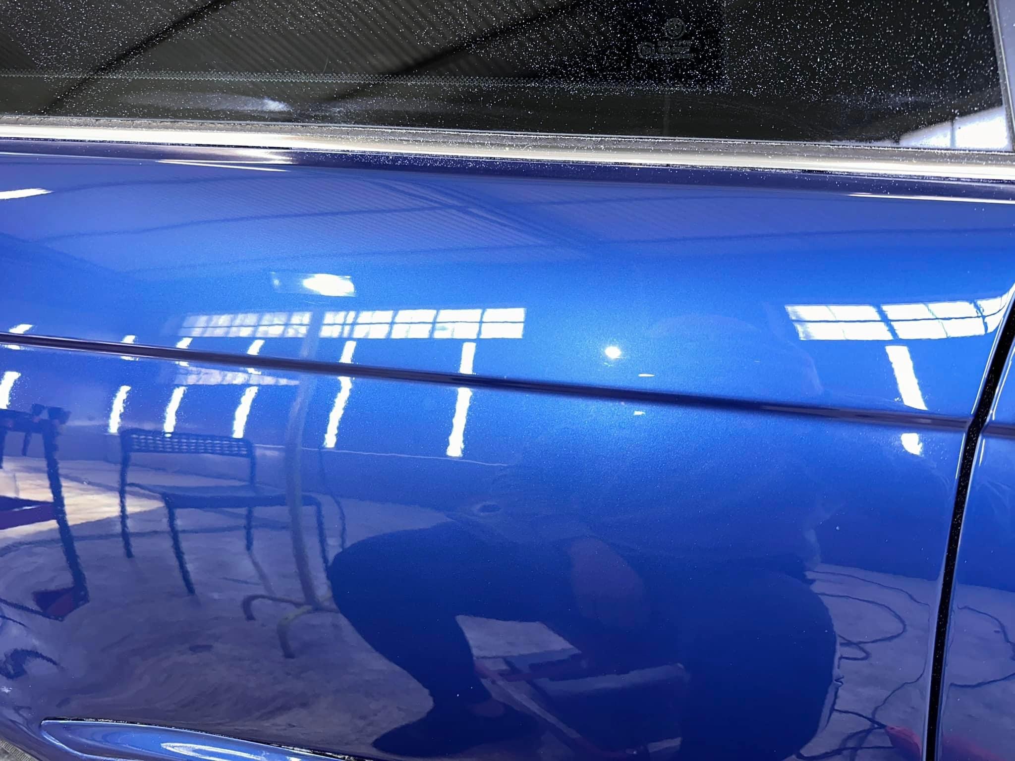 Błyszcząca, niebieska karoseria samochodu po polerowaniu, widoczne odbicie otoczenia, w tym krzesła i osoby.