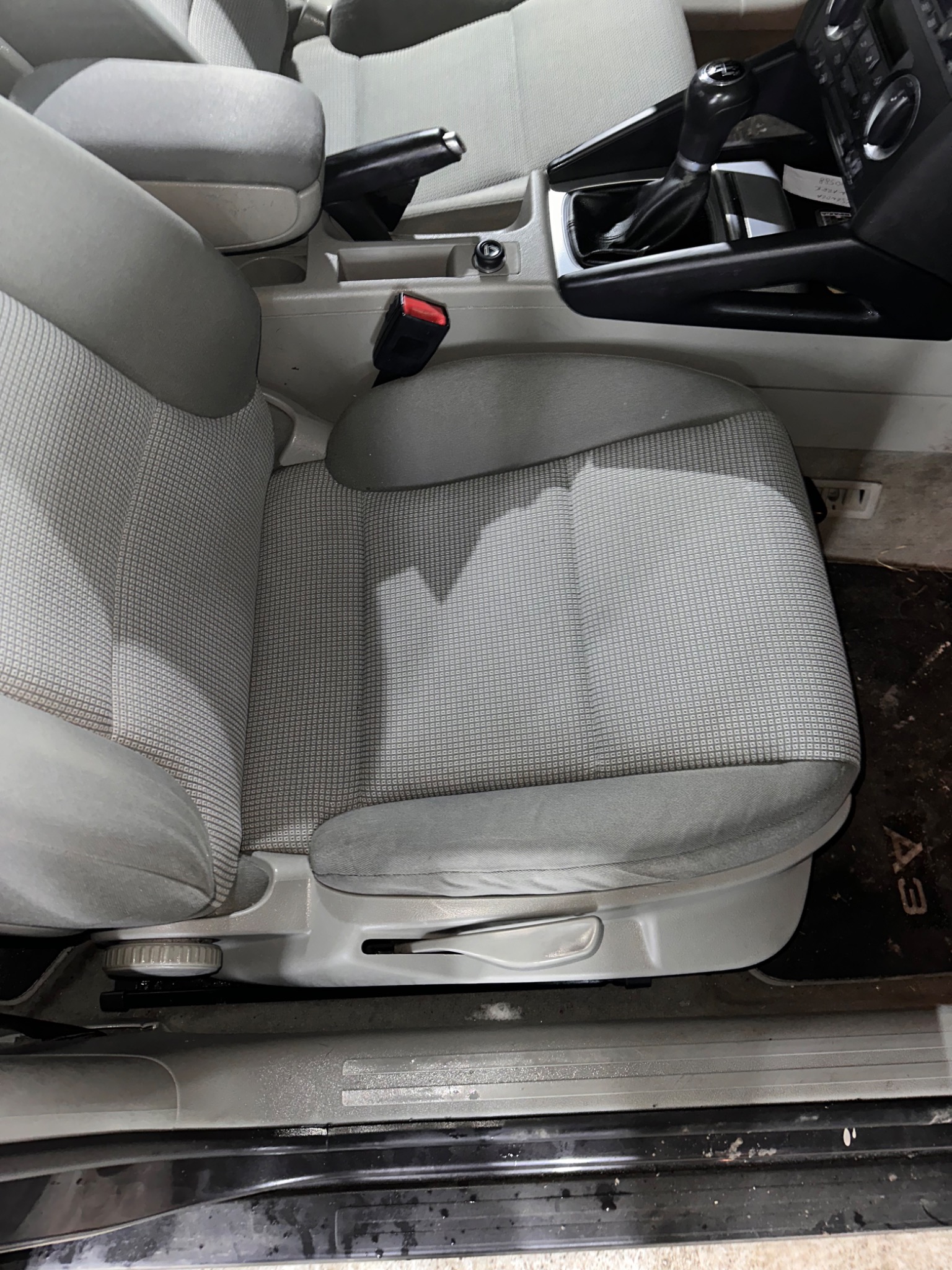 Szare, tekstylne siedzenie samochodowe z widocznym mechanizmem regulacji, z plamami na dywaniku pod siedzeniem.
