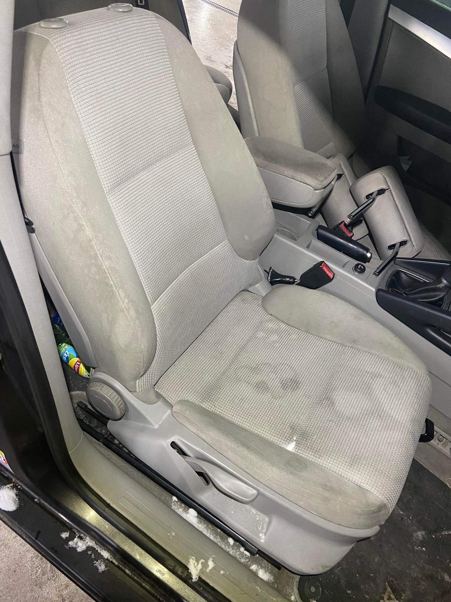 Brudny, jasnoszary fotel samochodowy z plamami i okruchami, widoczna butelka z napojem w szczelinie, otwarte drzwi auta.