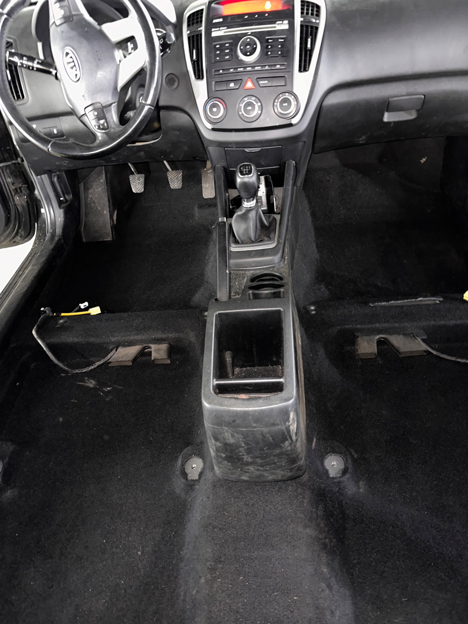 Wnętrze samochodu Kia Ceed z widoczną czarną wykładziną podłogową przed czyszczeniem, z odłączonymi przewodami i elementami montażowymi.