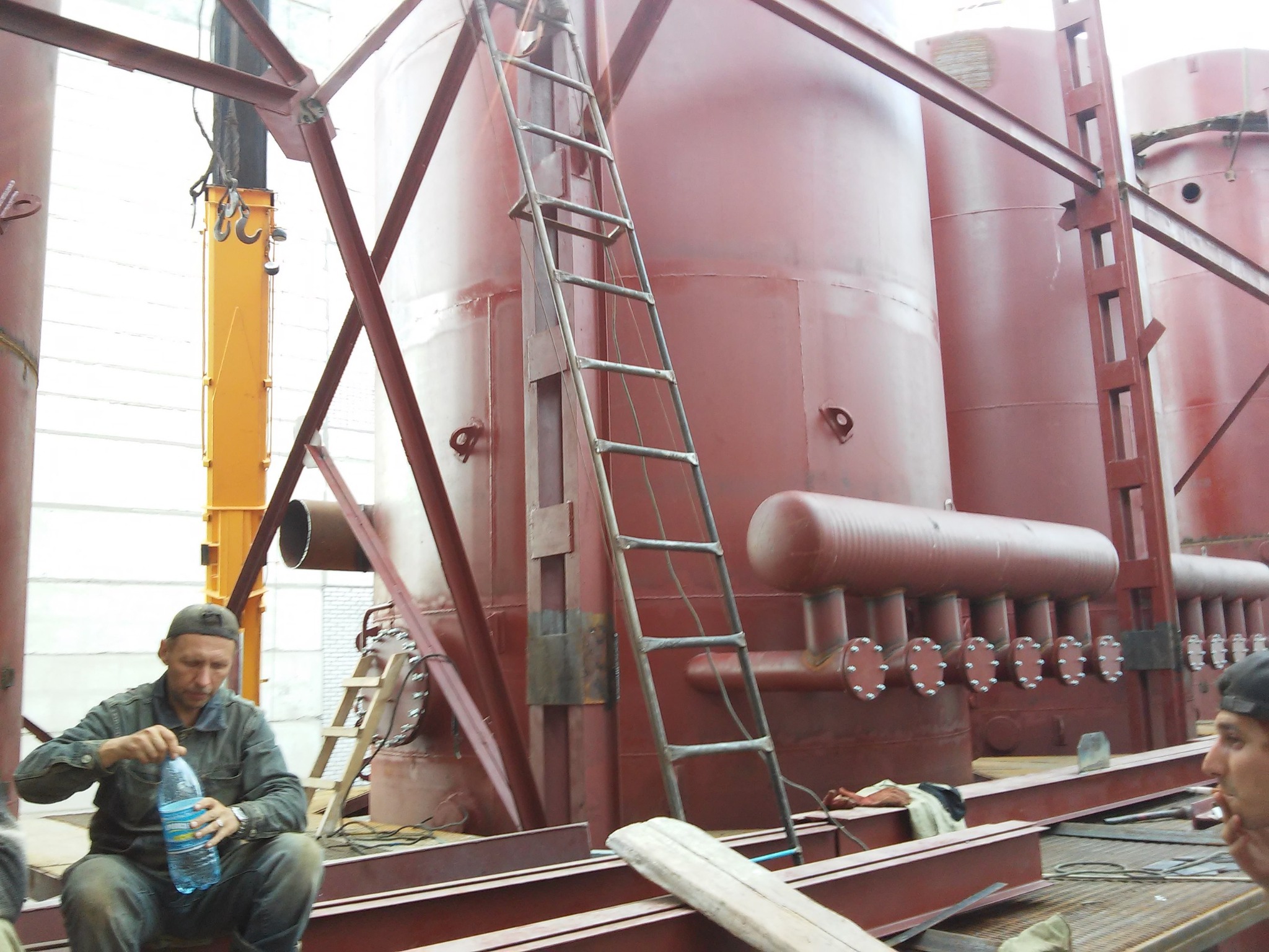 Montaż przemysłowych zbiorników stalowych w hali, widoczny pracownik otwierający butelkę wody, rusztowania i suwnica.