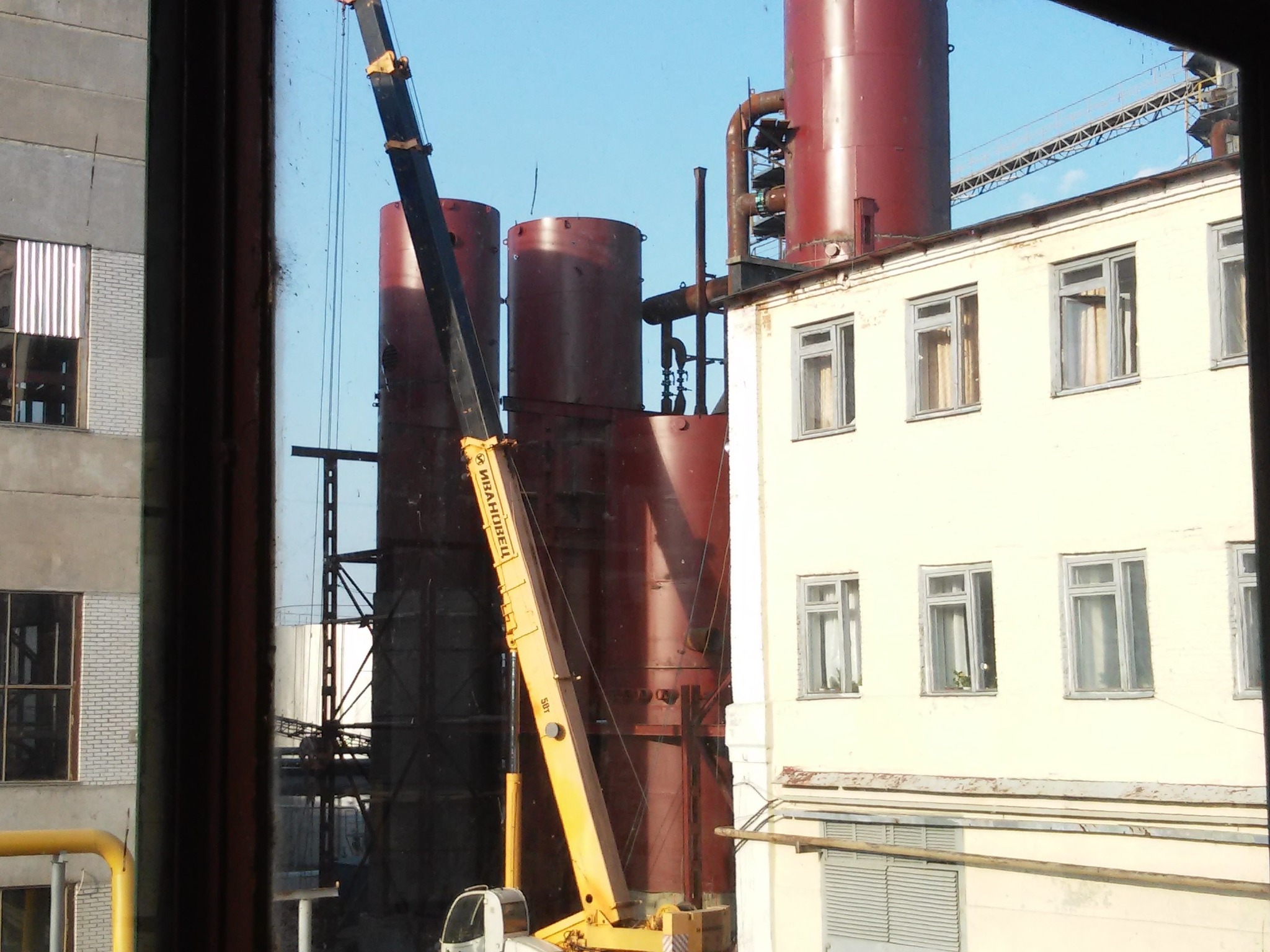 Montaż konstrukcji stalowej w Bydgoszczy z użyciem dźwigu, widok z okna na budynek przemysłowy i zbiorniki.