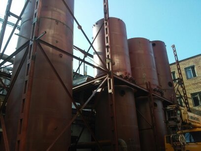 Zewnętrzne zbiorniki przemysłowe wsparte na stalowej konstrukcji kratownicowej, widoczne elementy instalacji i fragment budynku w tle.