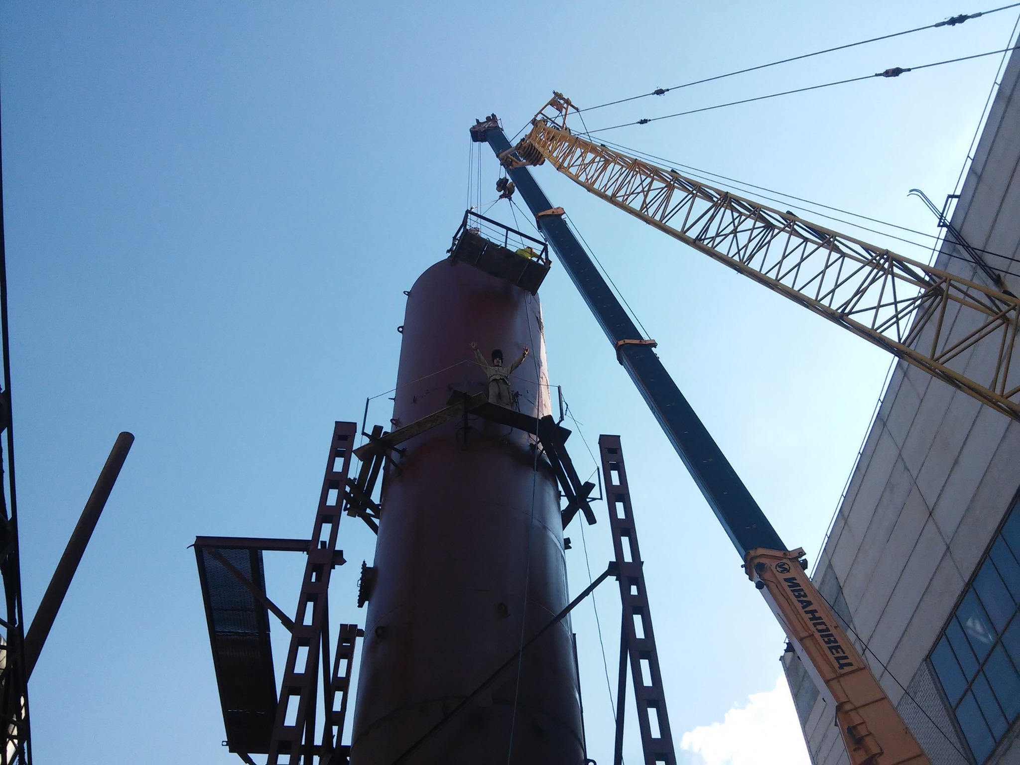 Montaż pionowego zbiornika przemysłowego przy użyciu dźwigu, pracownik na szczycie z podniesionymi rękami, elementy konstrukcji stalowej w tle.