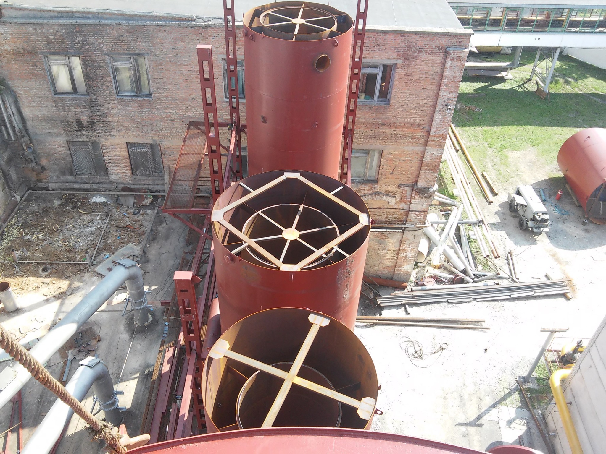 Widok z góry na trzy cylindryczne elementy konstrukcji stalowej w kolorze czerwono-brązowym, ustawione jeden na drugim w pionowym szybie. Elementy posiadają wewnętrzne wzmocnienia. W tle budynek...