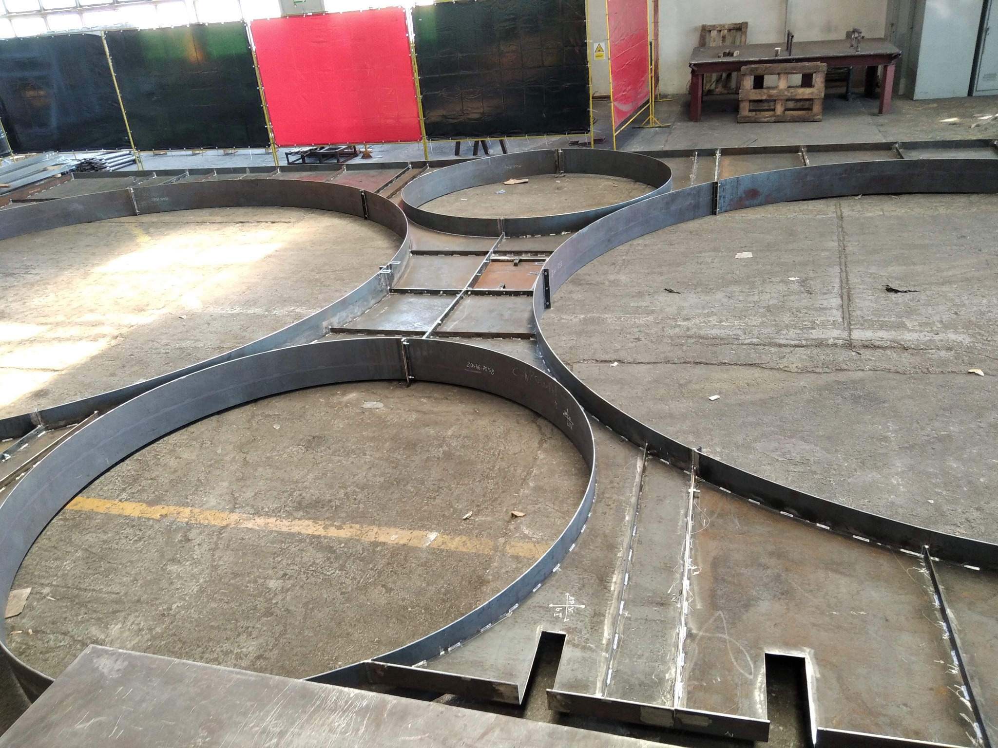 Metalowe elementy konstrukcyjne w kształcie okręgów i prostych odcinków, połączone spawami, leżące na betonowej podłodze hali produkcyjnej, z widocznymi żółtymi liniami i osłonami spawalniczymi w tle.
