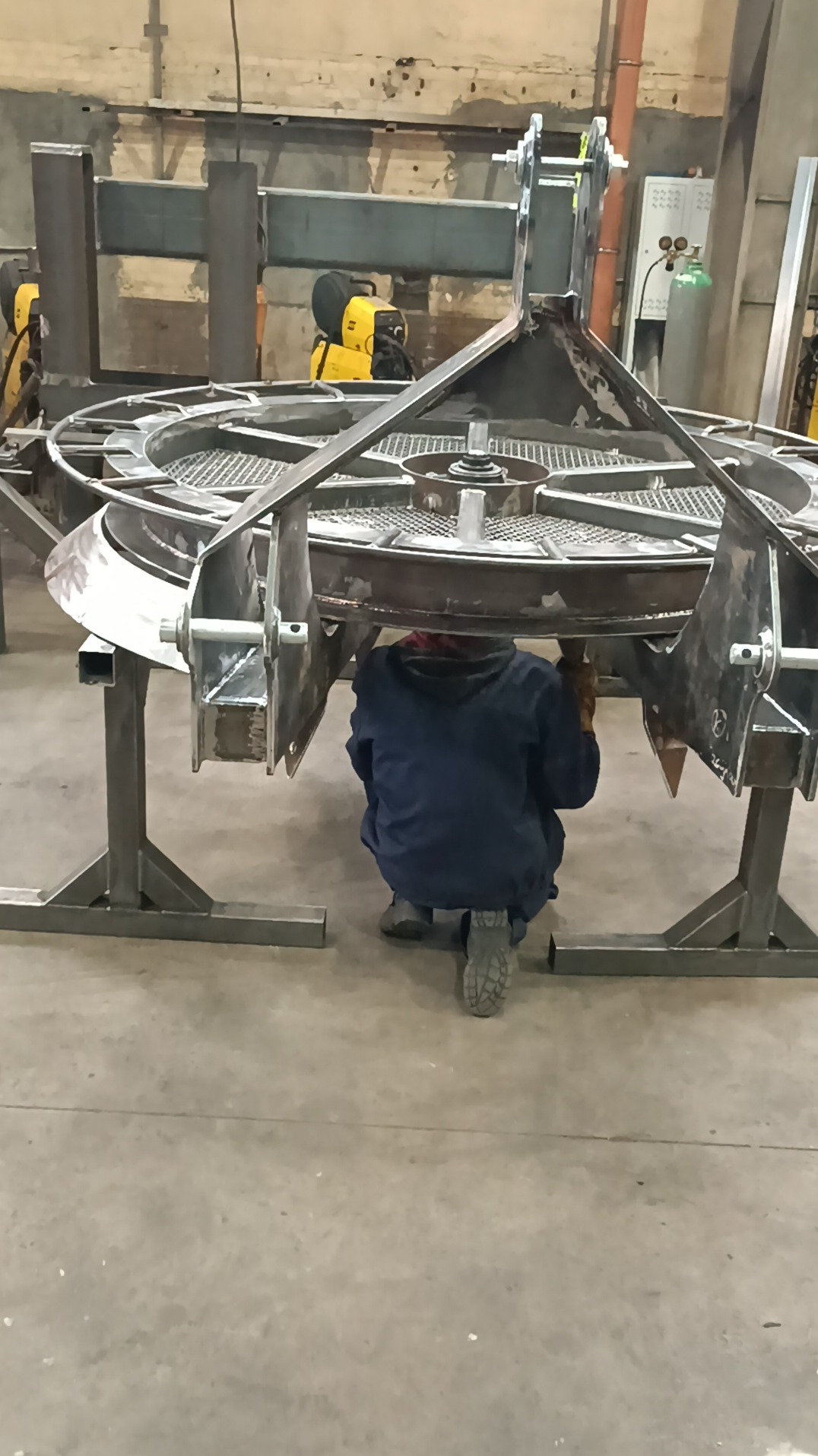 Pracownik w kombinezonie spawalniczym, w pozycji kucznej, pracuje pod dużą, okrągłą konstrukcją metalową z kratownicą, wspartą na stalowych nogach, w hali produkcyjnej.